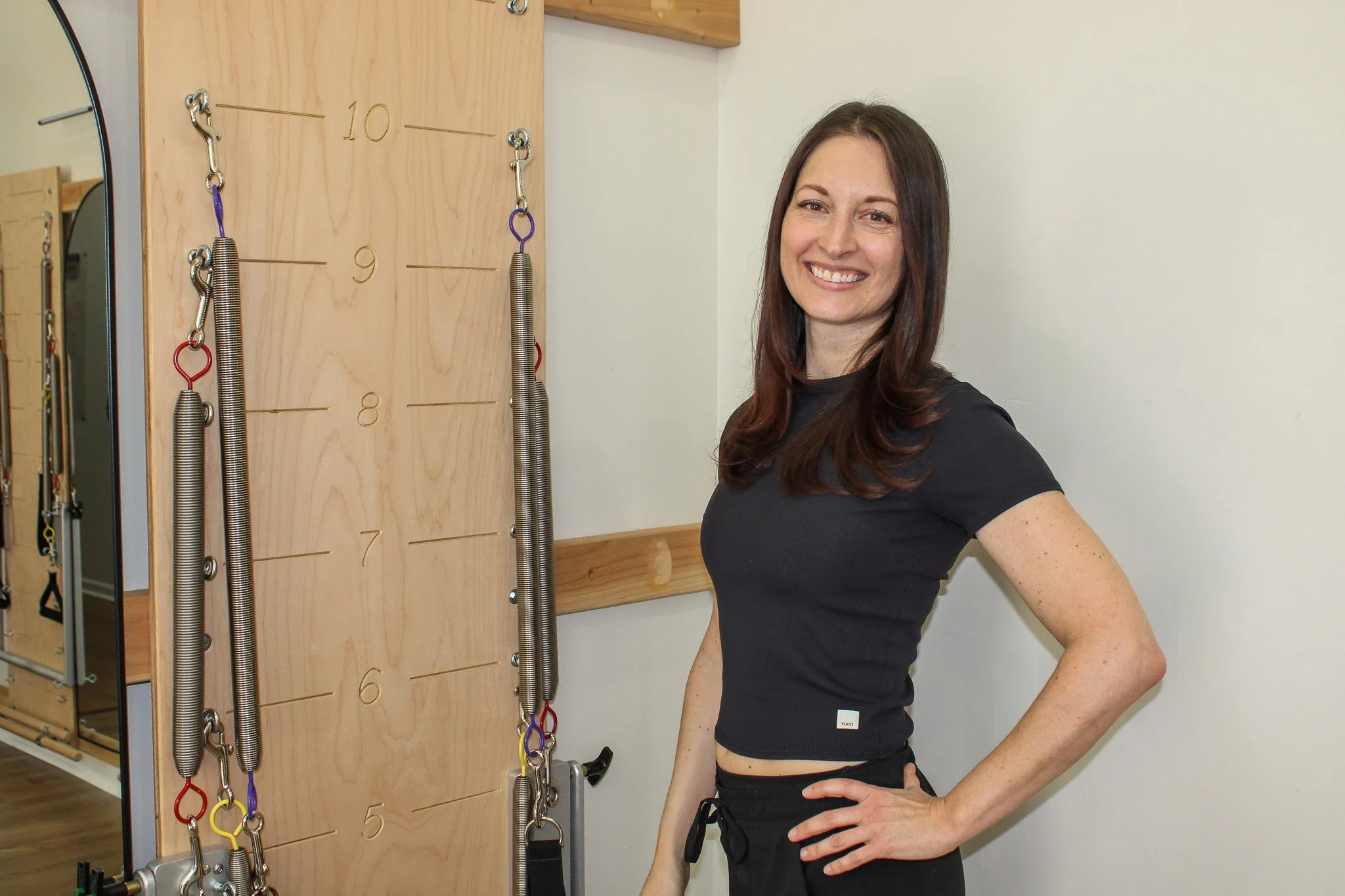 A woman with dark hair smiling, standing with her hand on her hip, next to a wooden wall with springs attached for exercise.