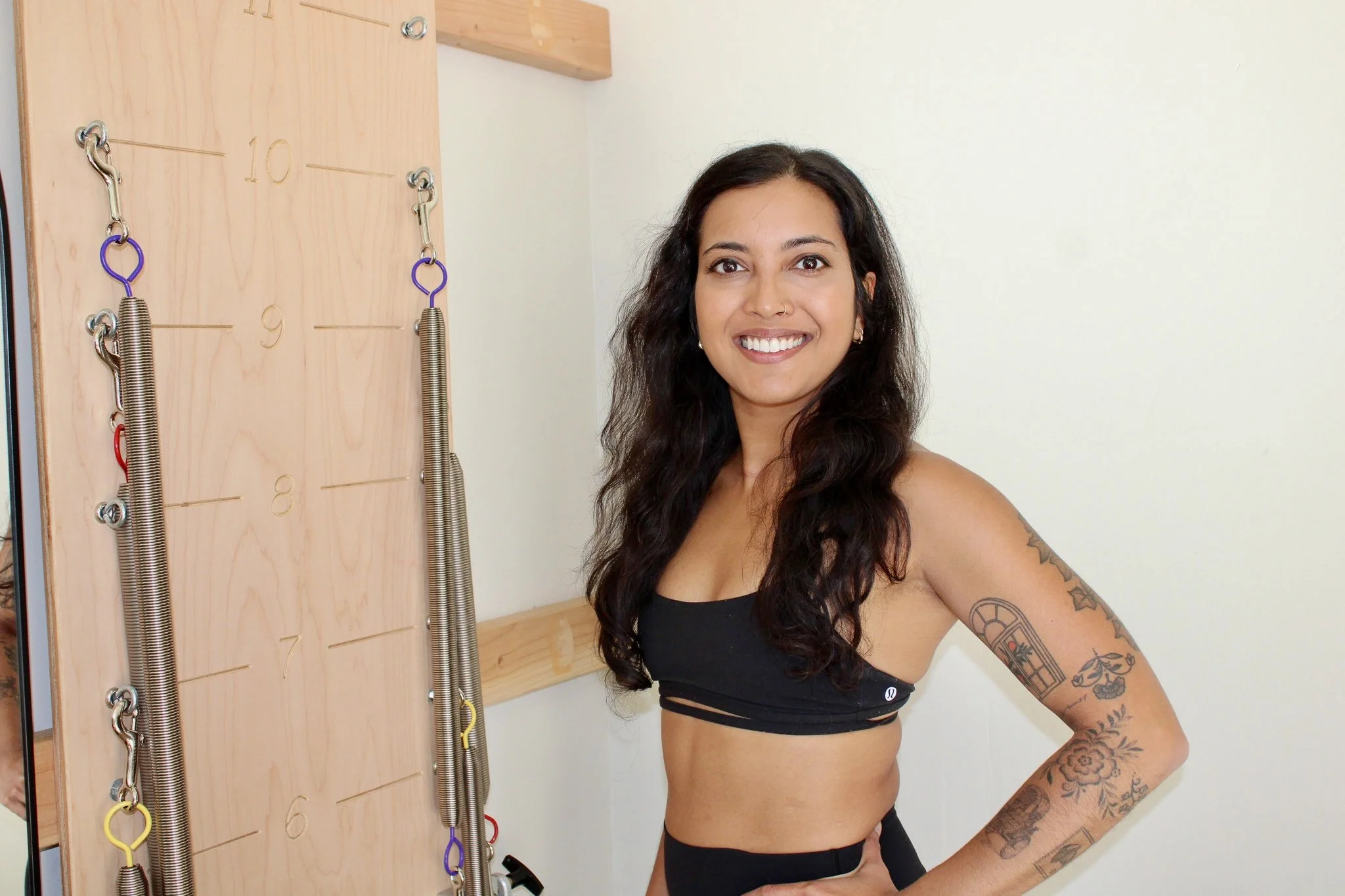 A smiling woman with long dark hair standing next to a wooden height measurement panel with a spring and purple hook attached.