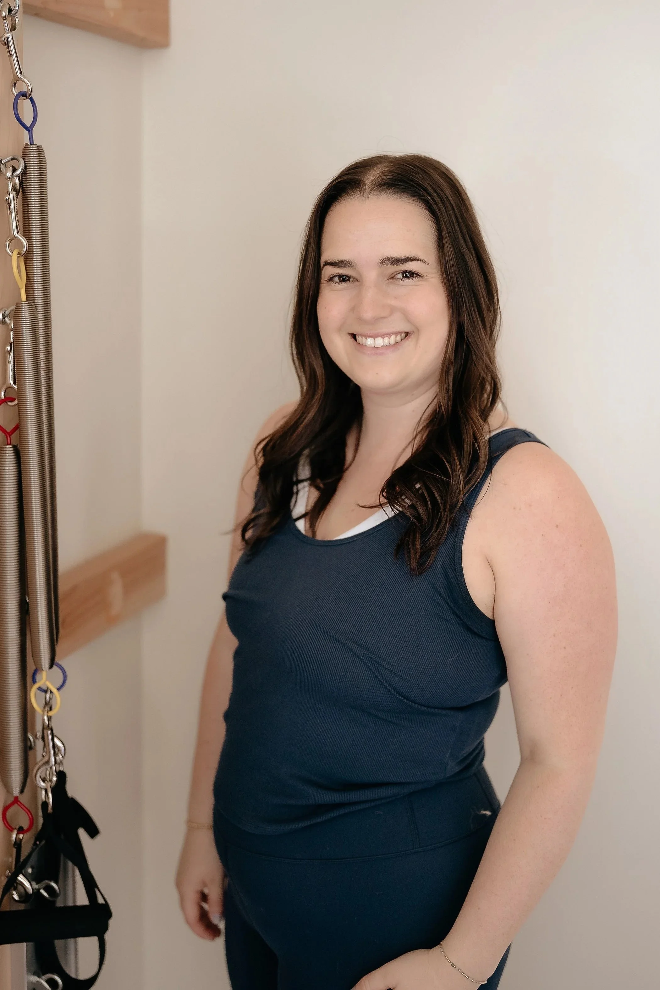 Smiling young woman with shoulder-length brown hair standing next to a large wooden agility or gymnastics training wall with numbered lines from 7 to 11 and metal hooks, springs, and a yellow and purple strap hanging from a hook.