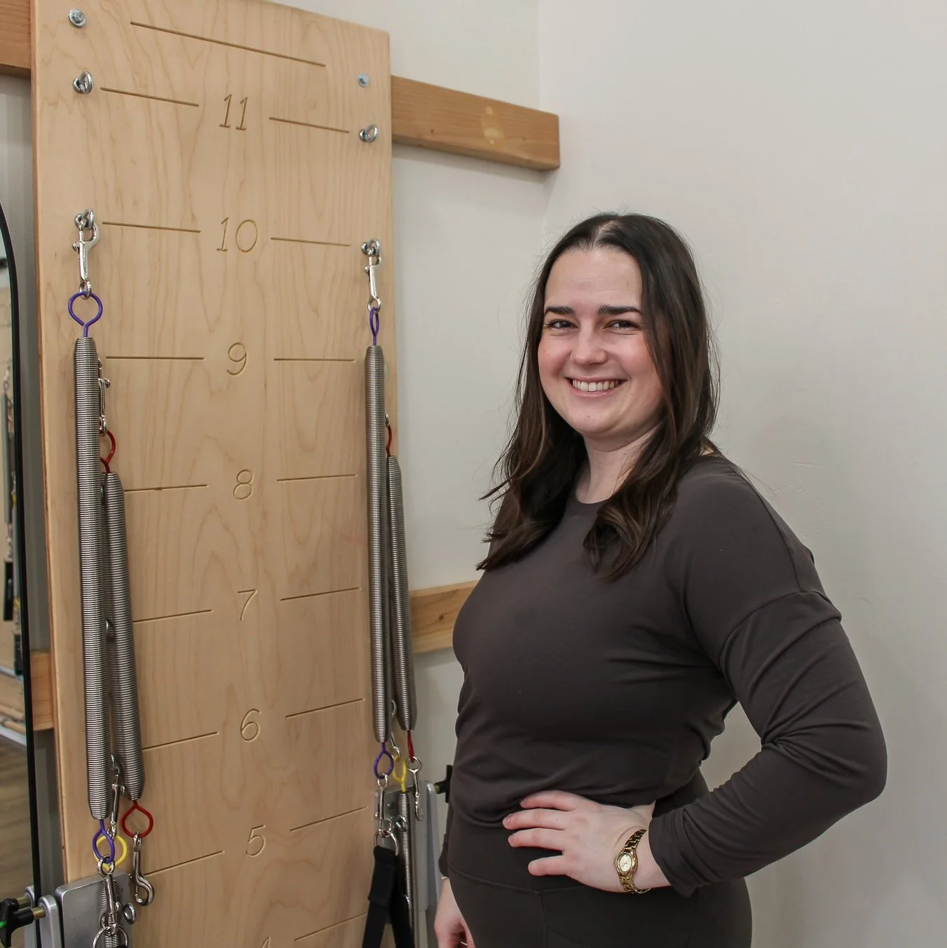 Smiling young woman with shoulder-length brown hair standing next to a large wooden agility or gymnastics training wall with numbered lines from 7 to 11 and metal hooks, springs, and a yellow and purple strap hanging from a hook.