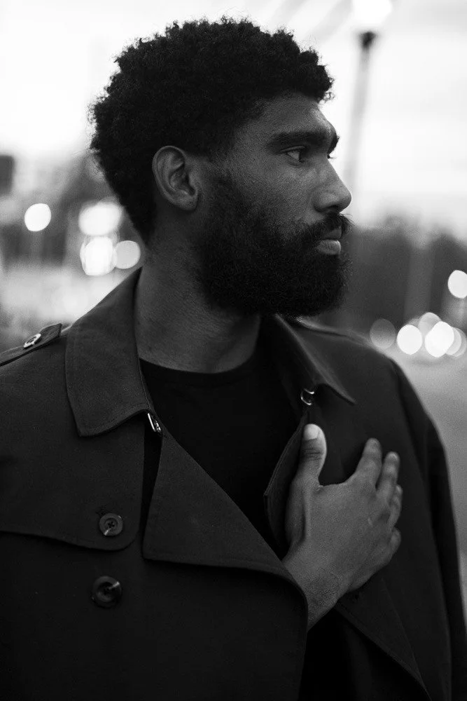 A black-and-white photo of a man with dark skin, curly hair, and a beard, standing outdoors with city lights blurred in the background. He is wearing a dark coat and has his right hand over his heart.
