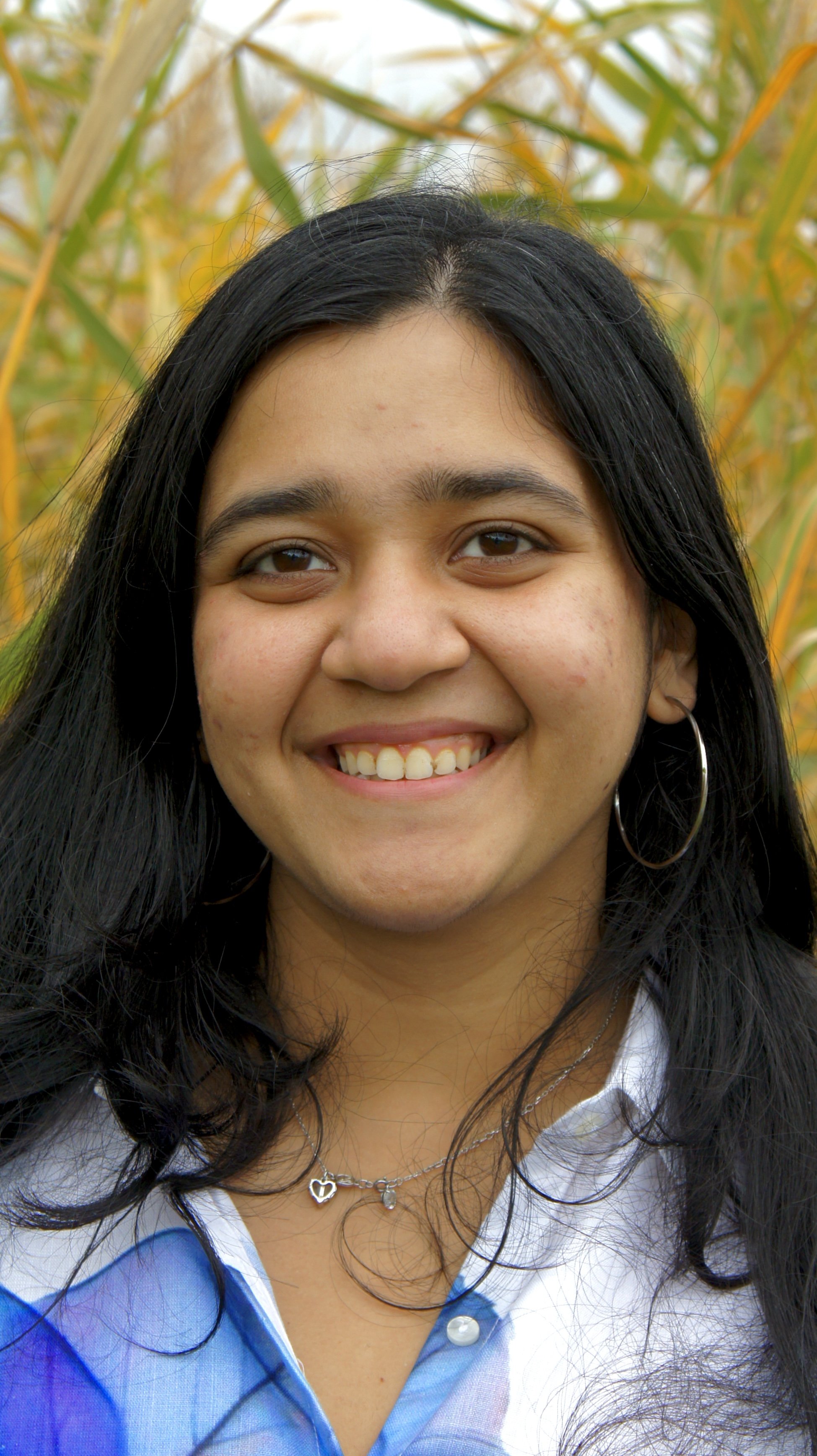 A young woman with long black hair and hoop earrings smiling outdoors, with tall grasses or plants in the background.