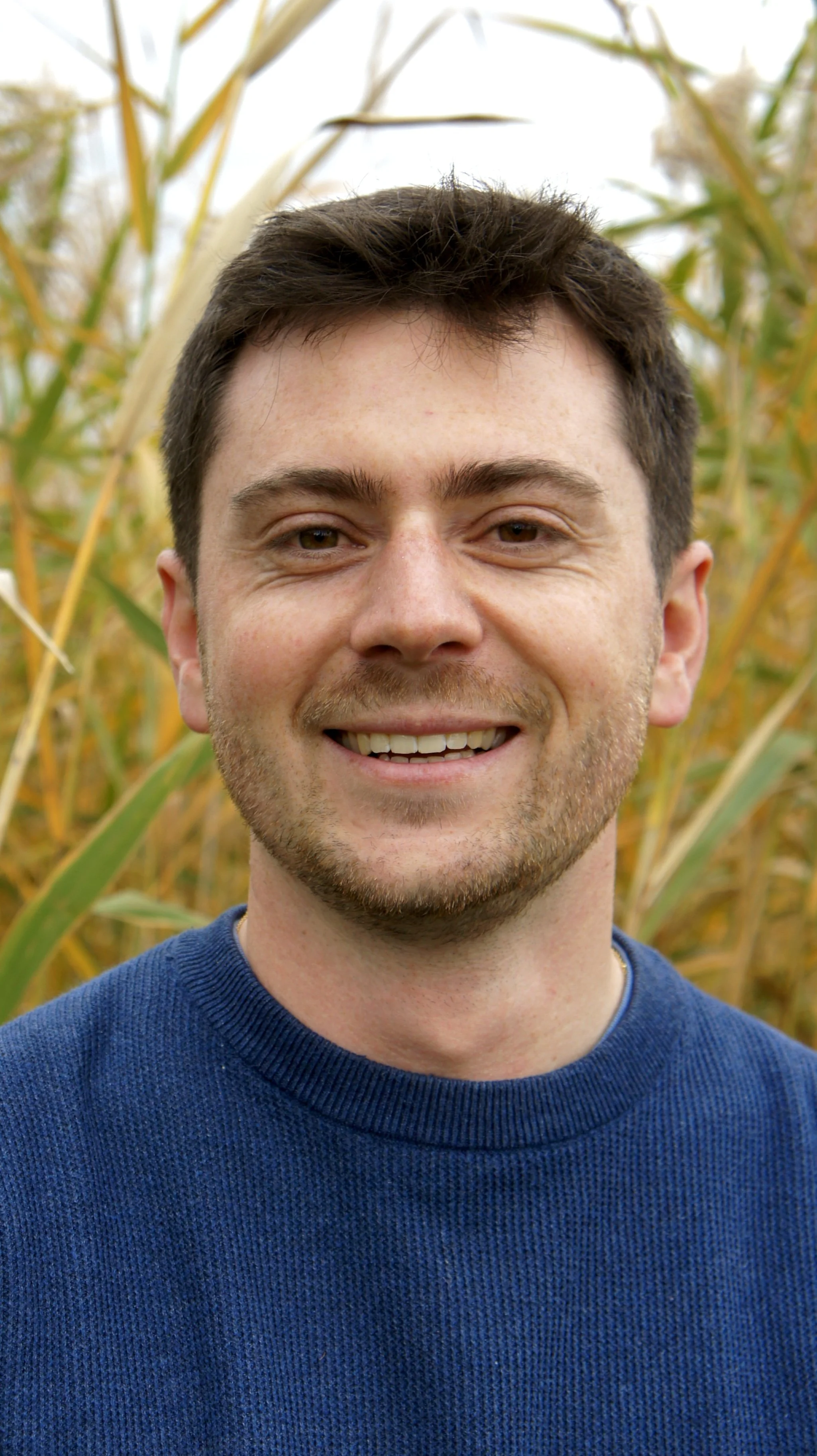 A smiling man with short dark hair and light stubble wearing a blue sweater, standing in a cornfield.