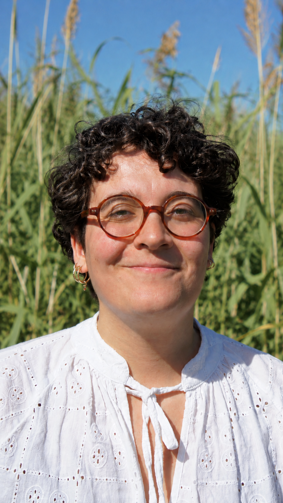 A smiling woman with short curly hair, wearing round glasses, earrings, and a white embroidered blouse, standing outdoors in a field of tall green crops under a clear blue sky.