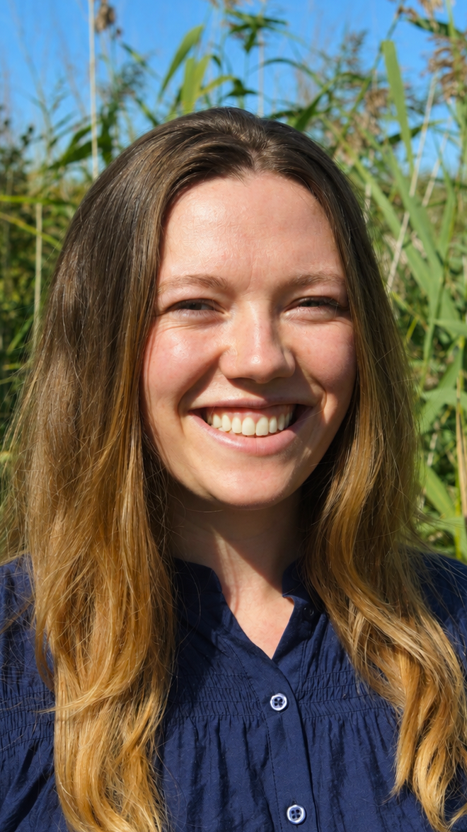 A woman with long, wavy light brown hair smiling outdoors with greenery and a blue sky in the background.