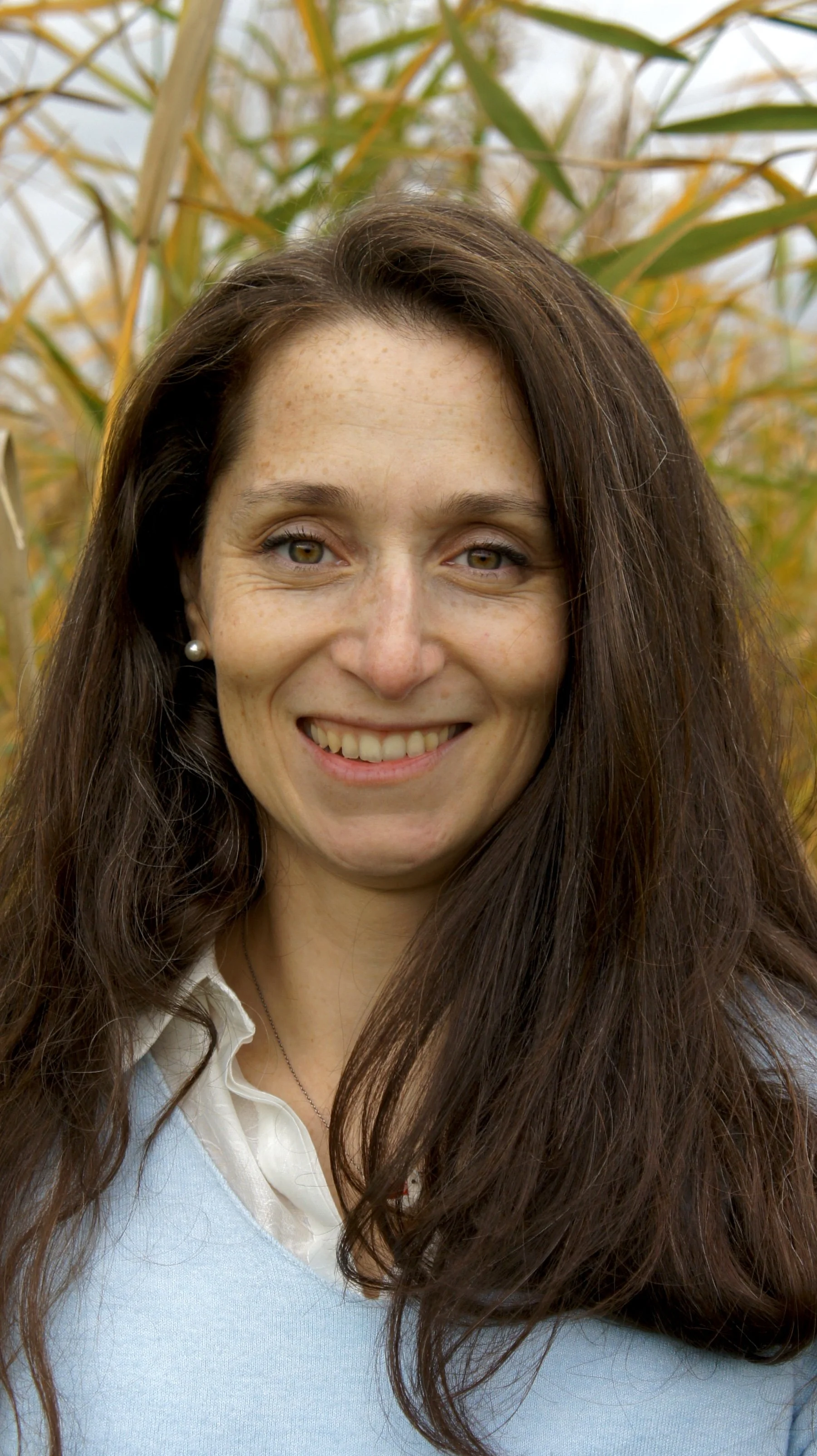 A woman with long brown hair smiling outdoors in a field with tall yellow grass.