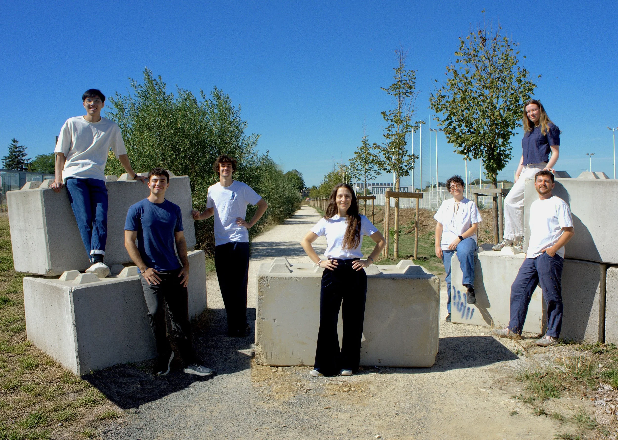 Group of seven young adults outdoors, some standing or sitting on large concrete blocks, with trees and a clear blue sky in the background.