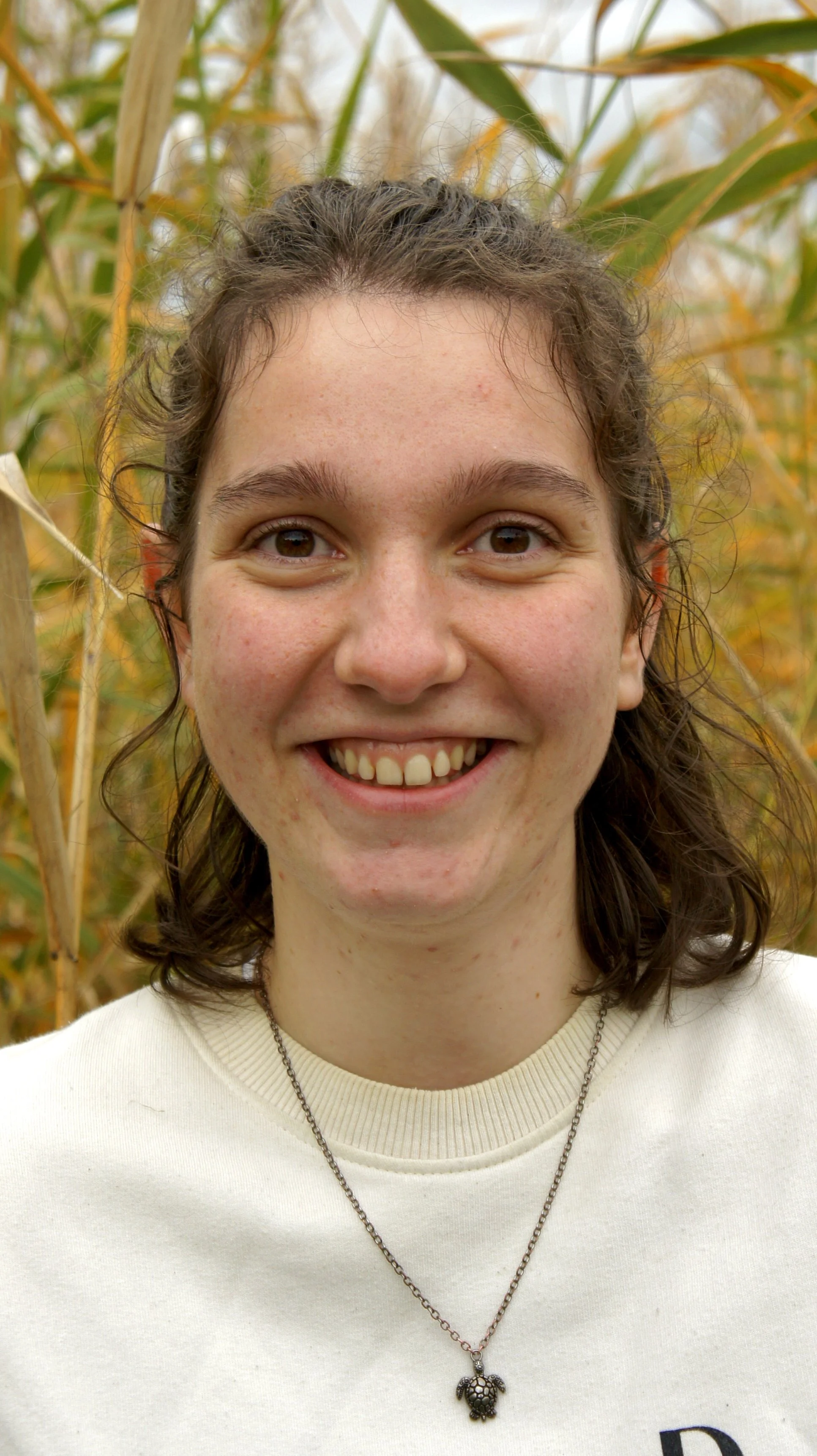 A young woman with curly brown hair, smiling, standing outdoors among tall yellowed corn stalks.