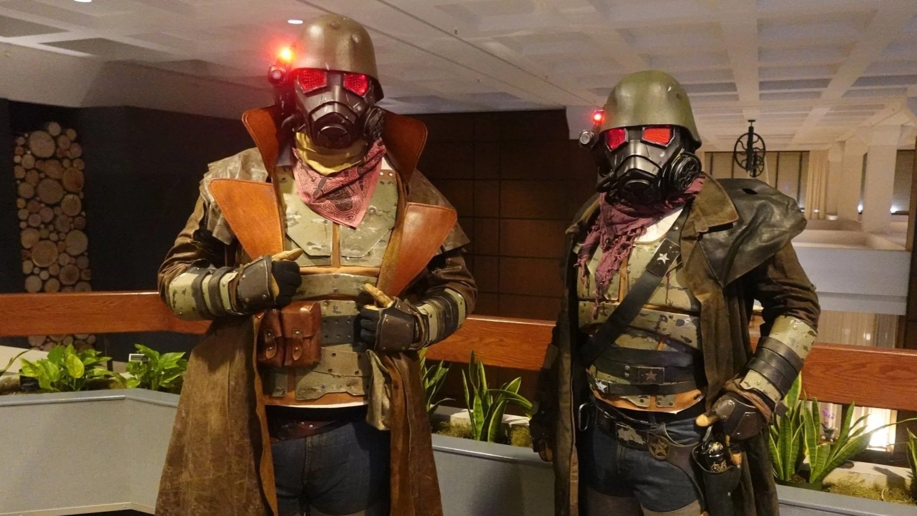Two individuals dressed as soldiers in tactical gear and gas masks with red glowing eyes, standing indoors near a wooden railing and green plants.