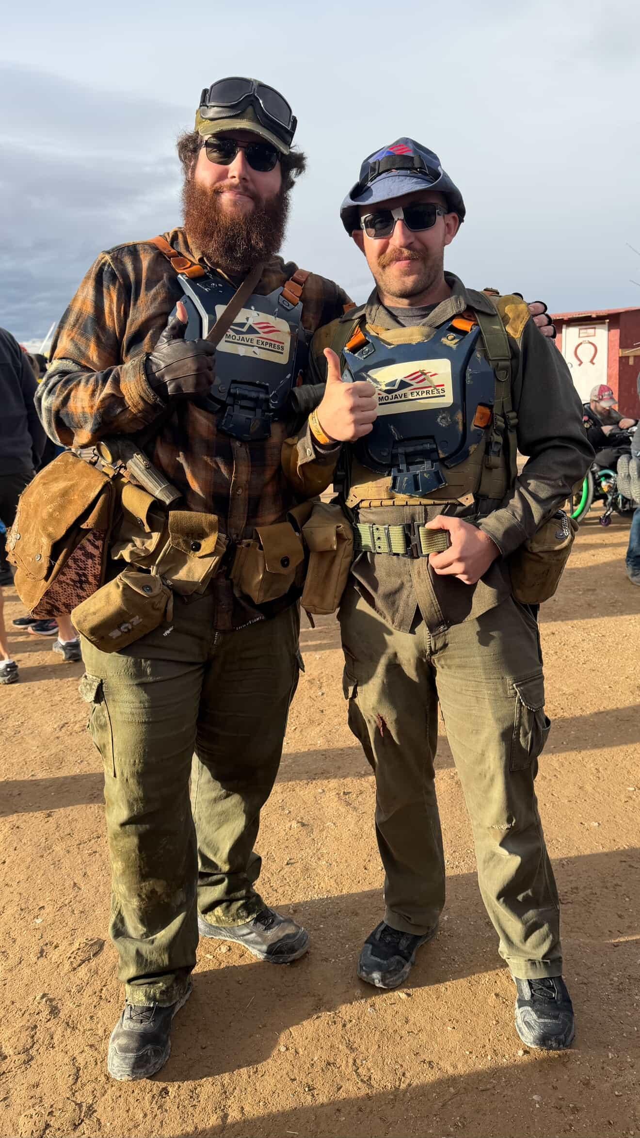 Two men wearing tactical gear and sunglasses, giving thumbs up at an outdoor event, with motorcycles and other people in the background.