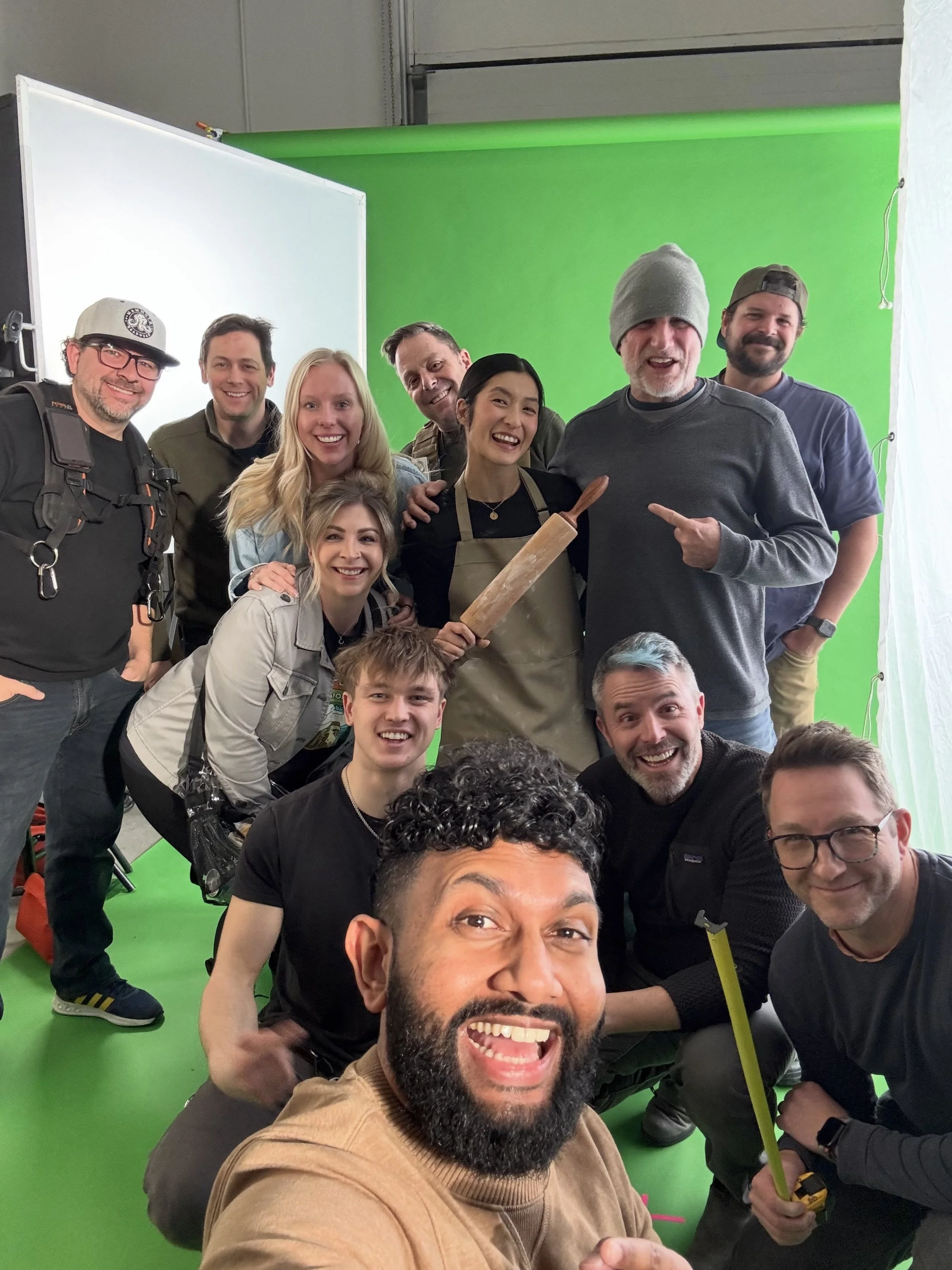 Group of people smiling and posing for a selfie in front of a green screen, some holding tools, in an indoor setting.