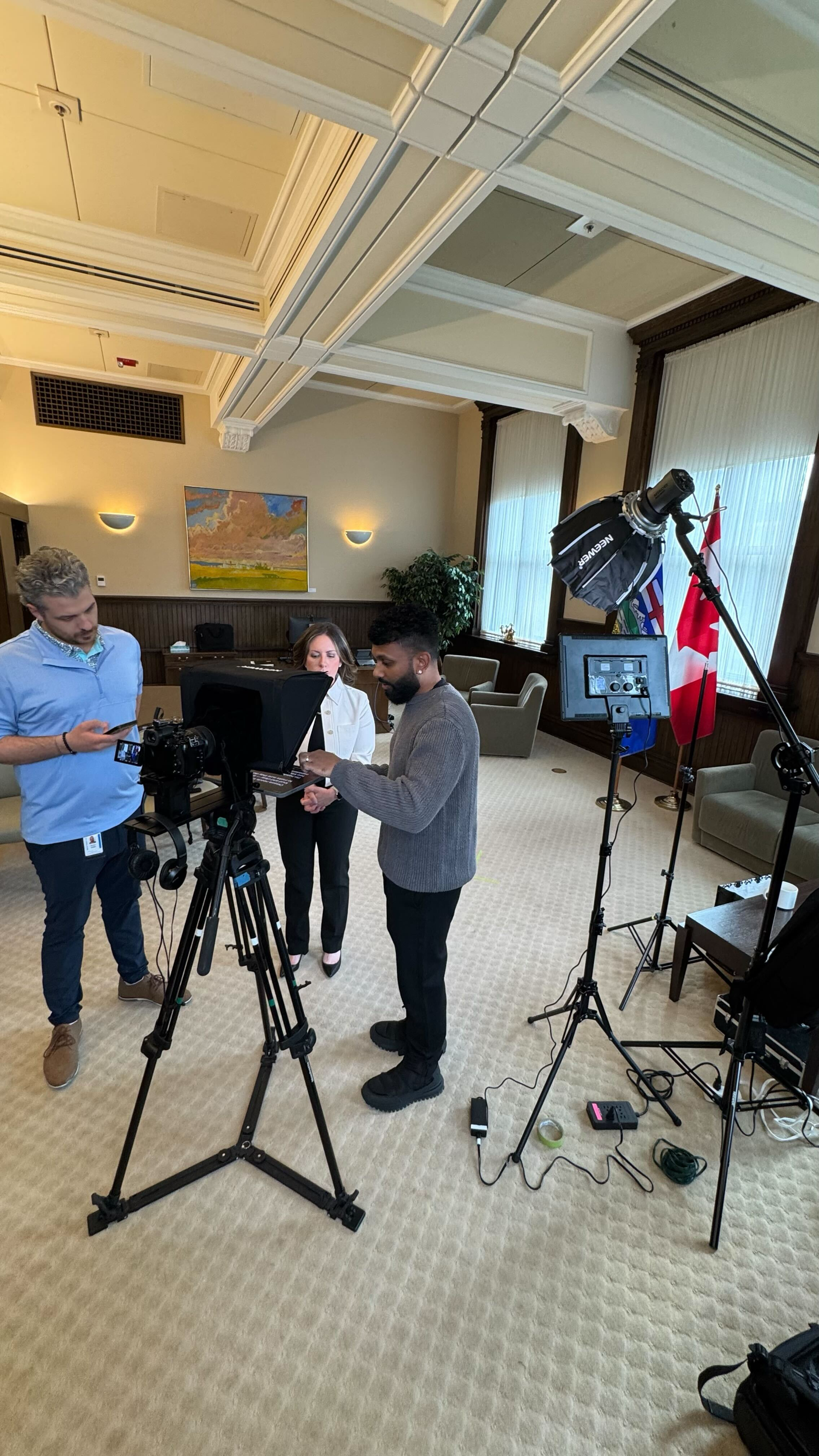 Three people working with camera and lighting equipment in a well-lit, formal room with beige carpet, wooden paneling, and large windows with curtains.