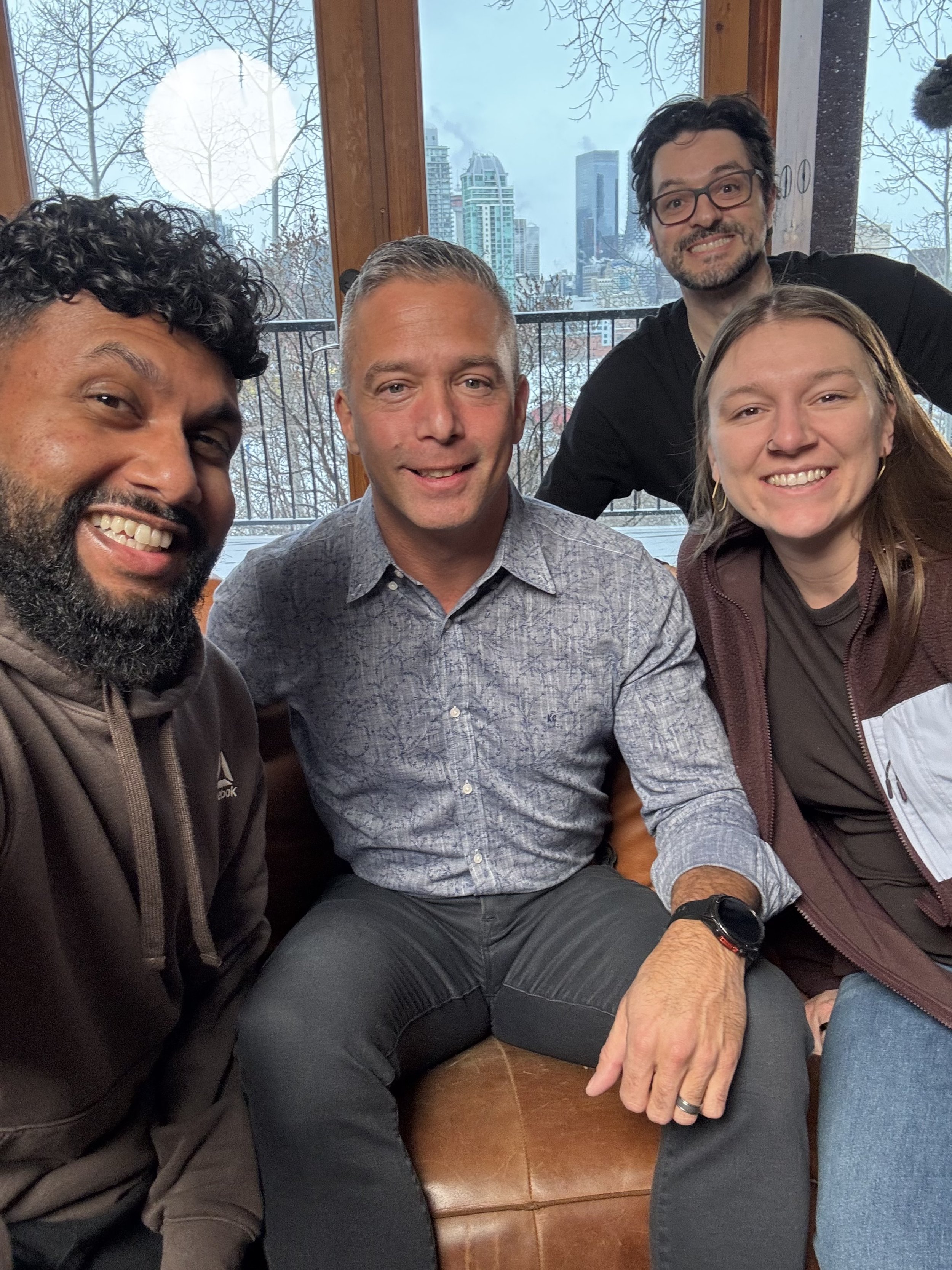 A group of five diverse people sitting and smiling together inside, with large windows in the background showing a cityscape on a cloudy day.