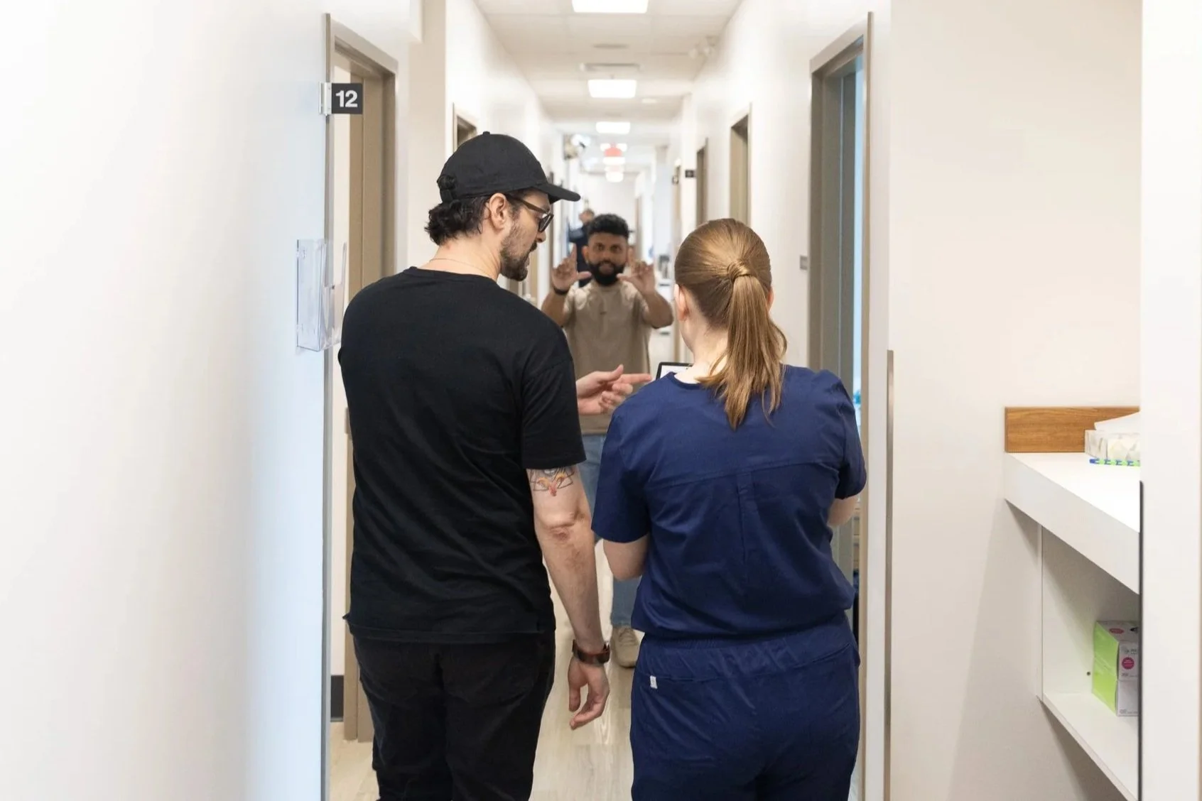 Two healthcare workers, one man and one woman, having a conversation in the hallway of a medical facility, with a man in the background making a peaceful gesture with his hands.
