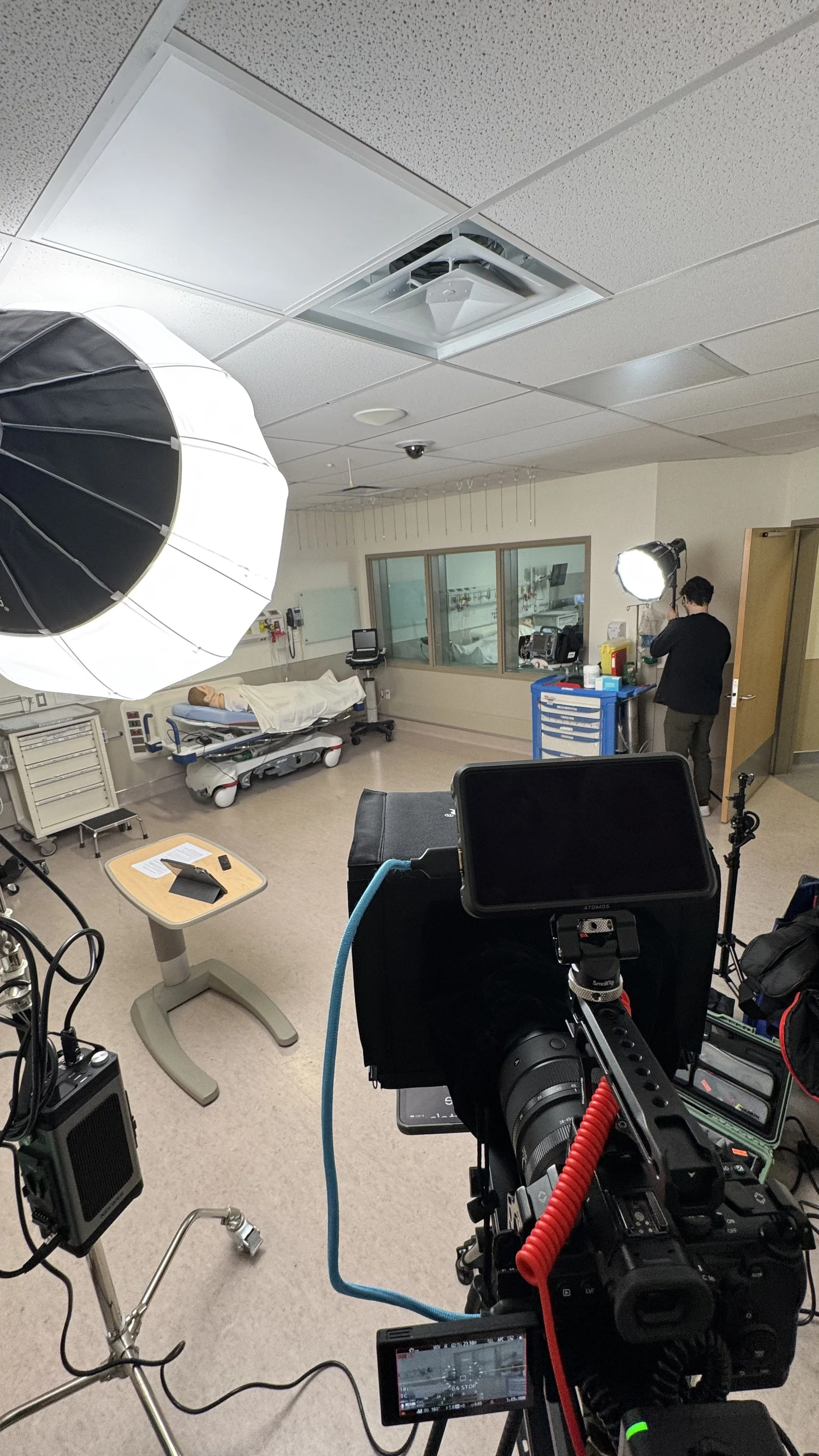 A hospital room with medical equipment, a patient on a bed, and a person adjusting a large studio light. Camera and lighting equipment are set up for filming or photography.