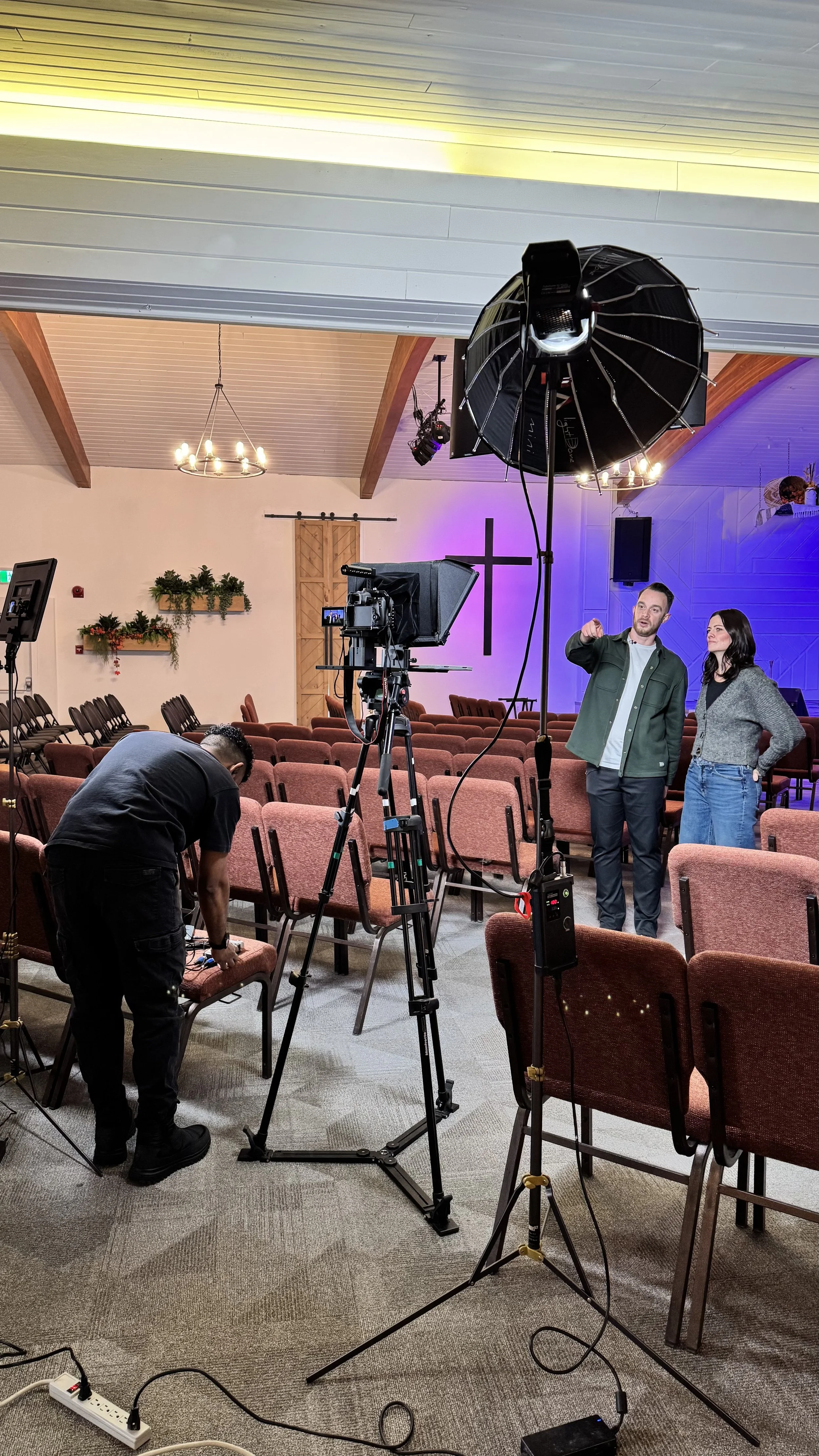 A film crew is setting up equipment inside a church with rows of red chairs, a central cross on the wall, and a hanging chandelier. Two people are talking, and another person is adjusting equipment.