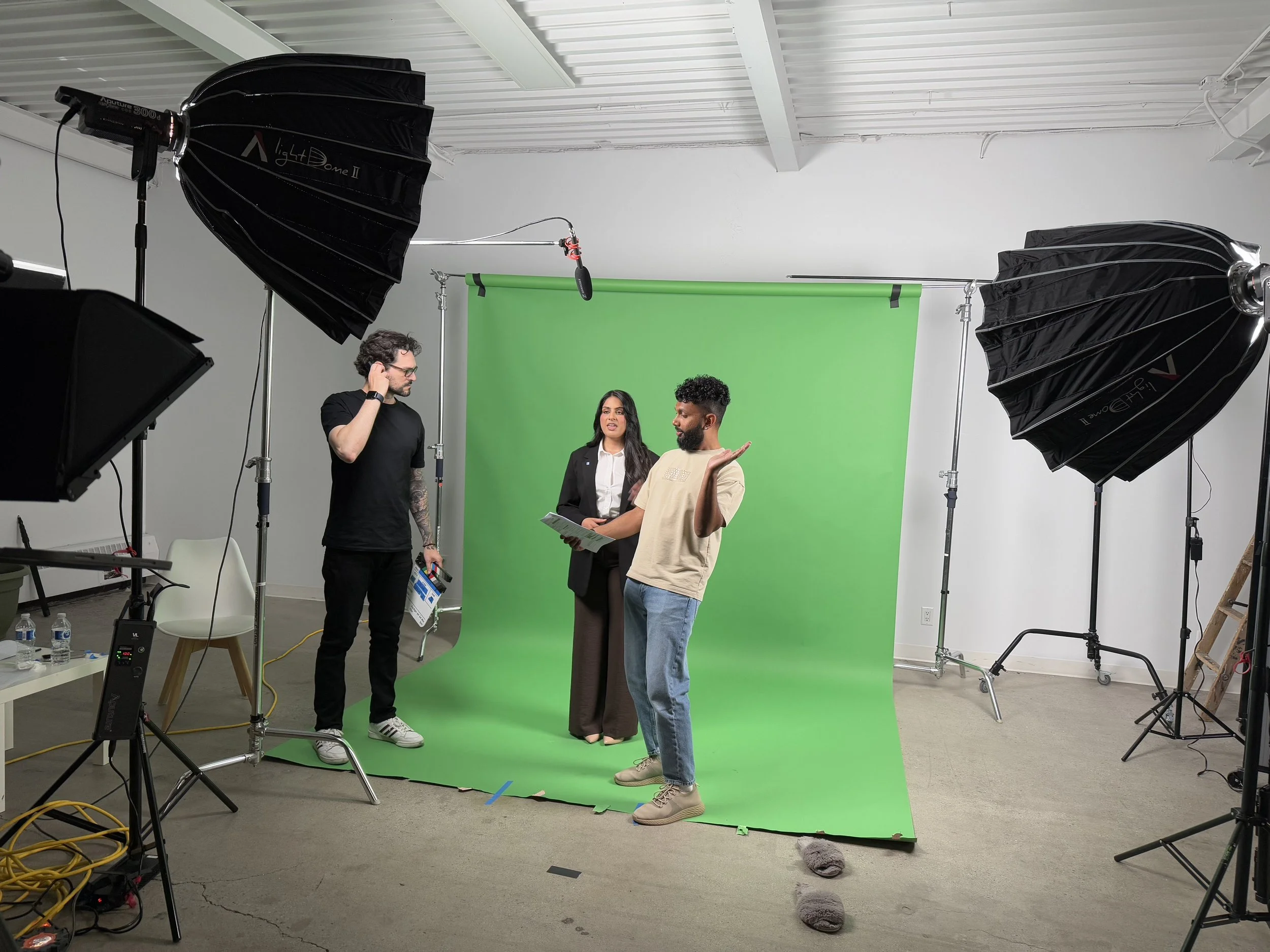 Three people in a photography studio with a green screen backdrop, large studio lights, and equipment visible.