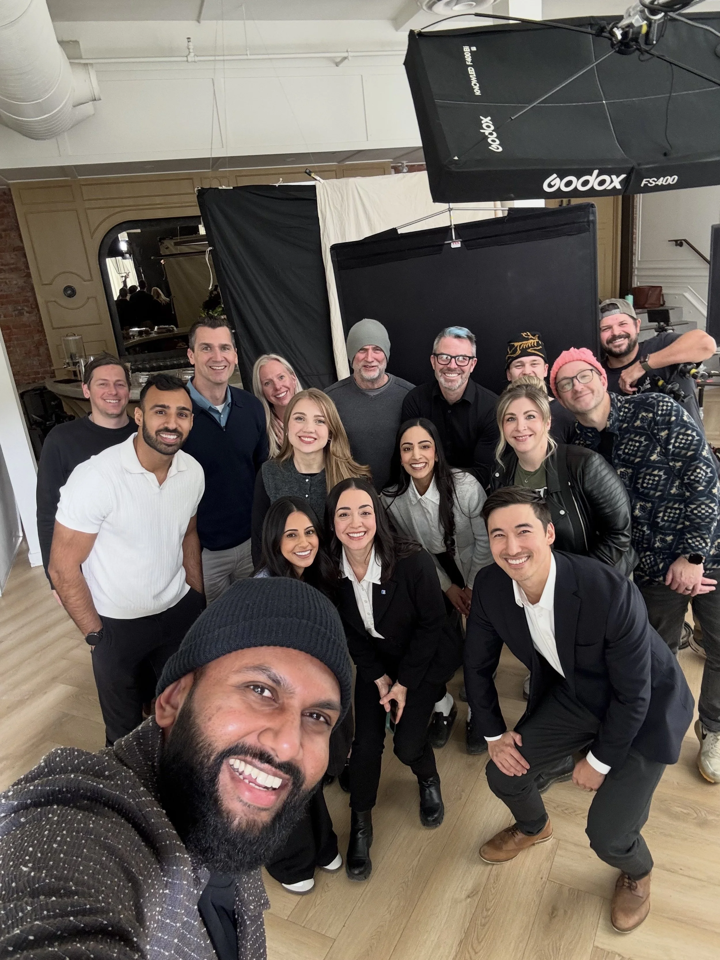 A diverse group of people smiling and posing for a selfie at an indoor event, with photography equipment, black curtains, and a mirror in the background.