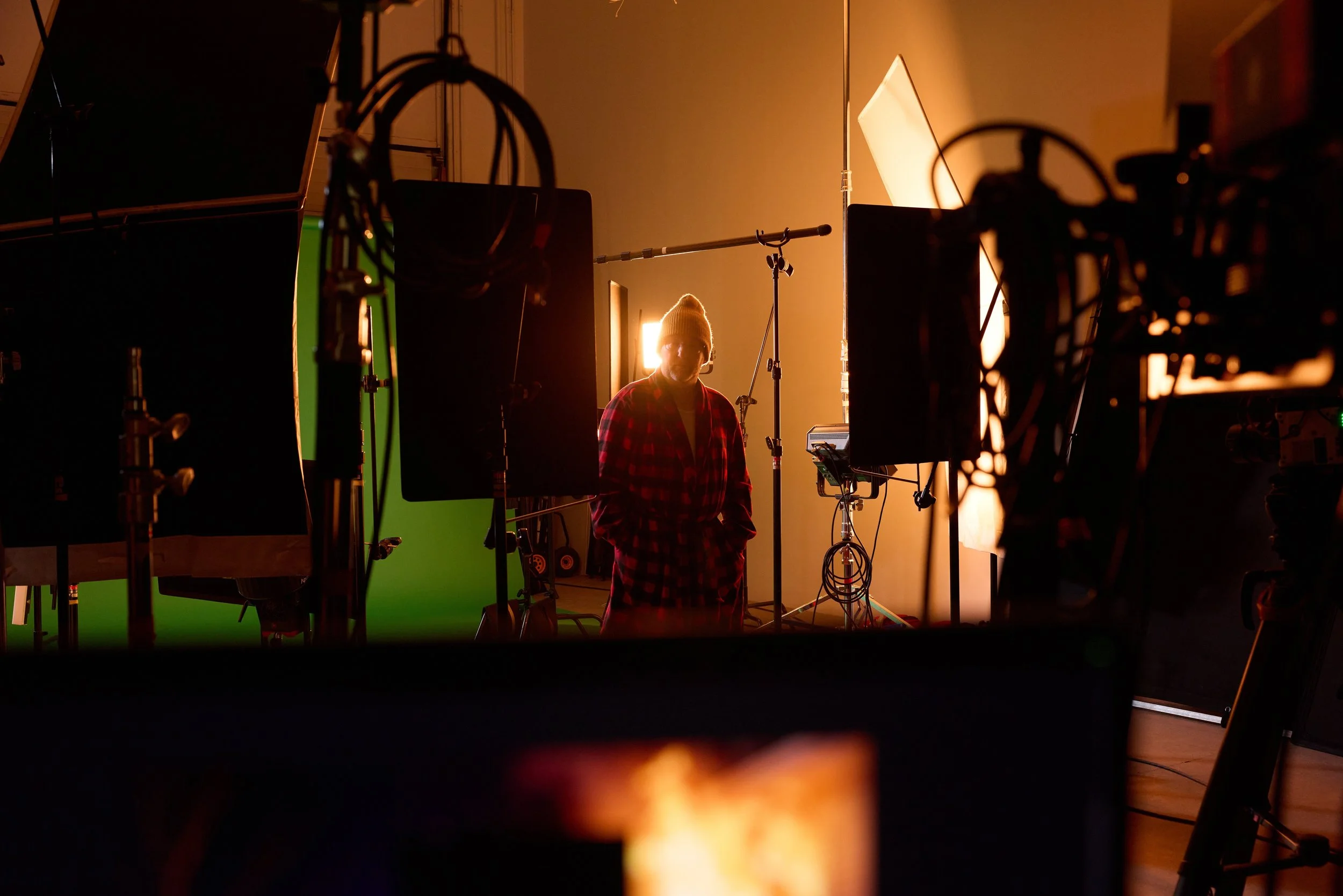 A person wearing a yellow beanie and red plaid shirt is on a film set surrounded by lighting equipment, partially illuminated by warm light, with green screen visible behind them.