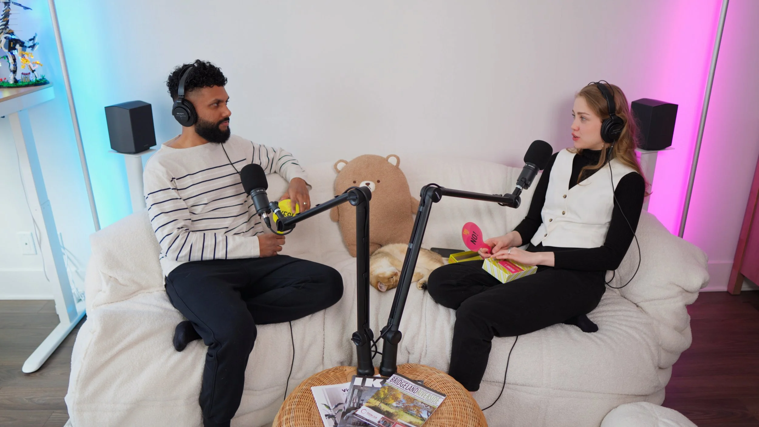 Two people, a man and a woman, are recording a podcast in a cozy room with led lights. They are sitting on a beige couch with a teddy bear and a sleeping dog between them. Each has a microphone and wears headphones.