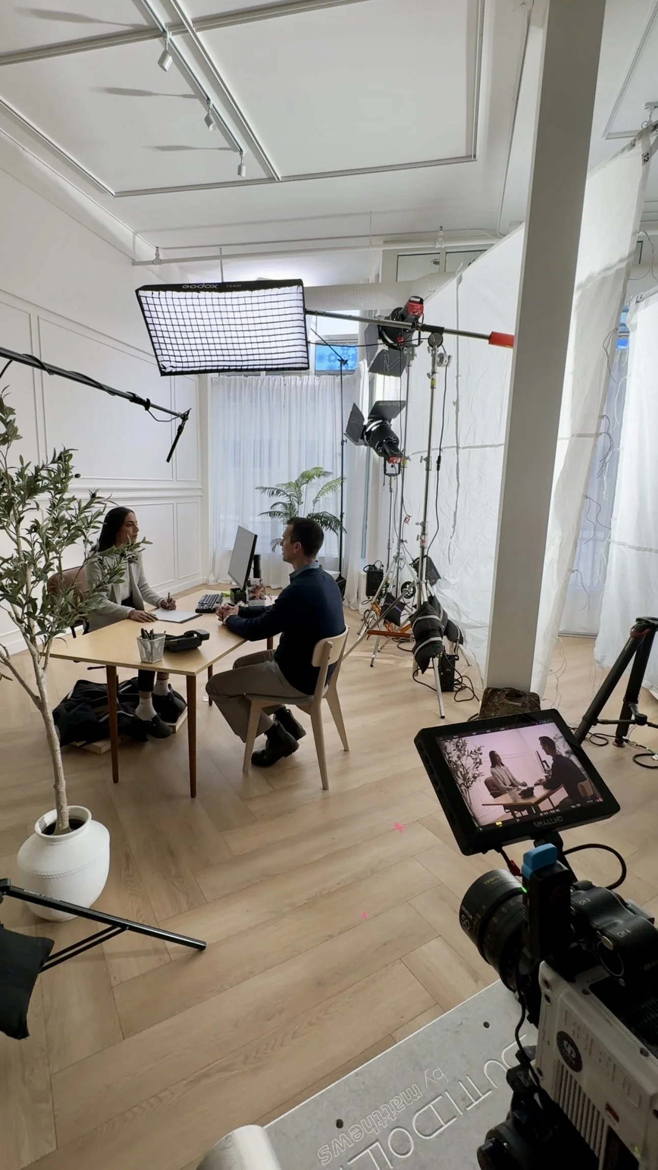 A video shoot or interview scene with two people seated at a table, filming equipment and lighting setup in a bright, white room with wooden flooring.