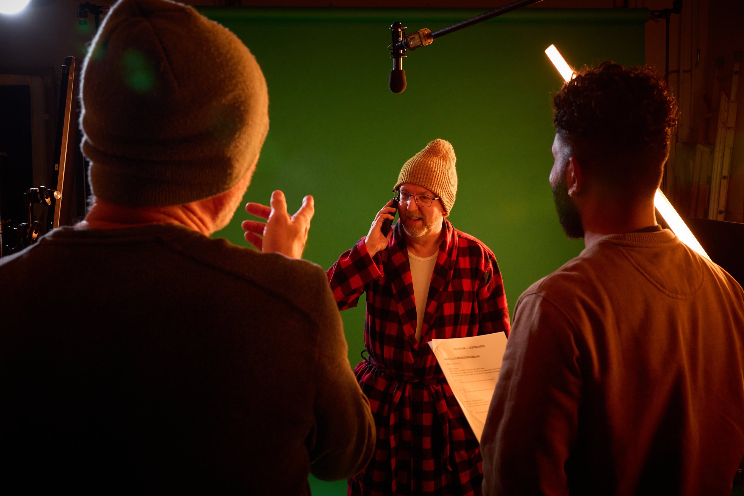 Behind the scenes of a film production, a man with glasses and a beige beanie is talking on the phone and holding a script, surrounded by two others in a dimly lit studio with a green screen backdrop, professional lighting, and a microphone overhead.