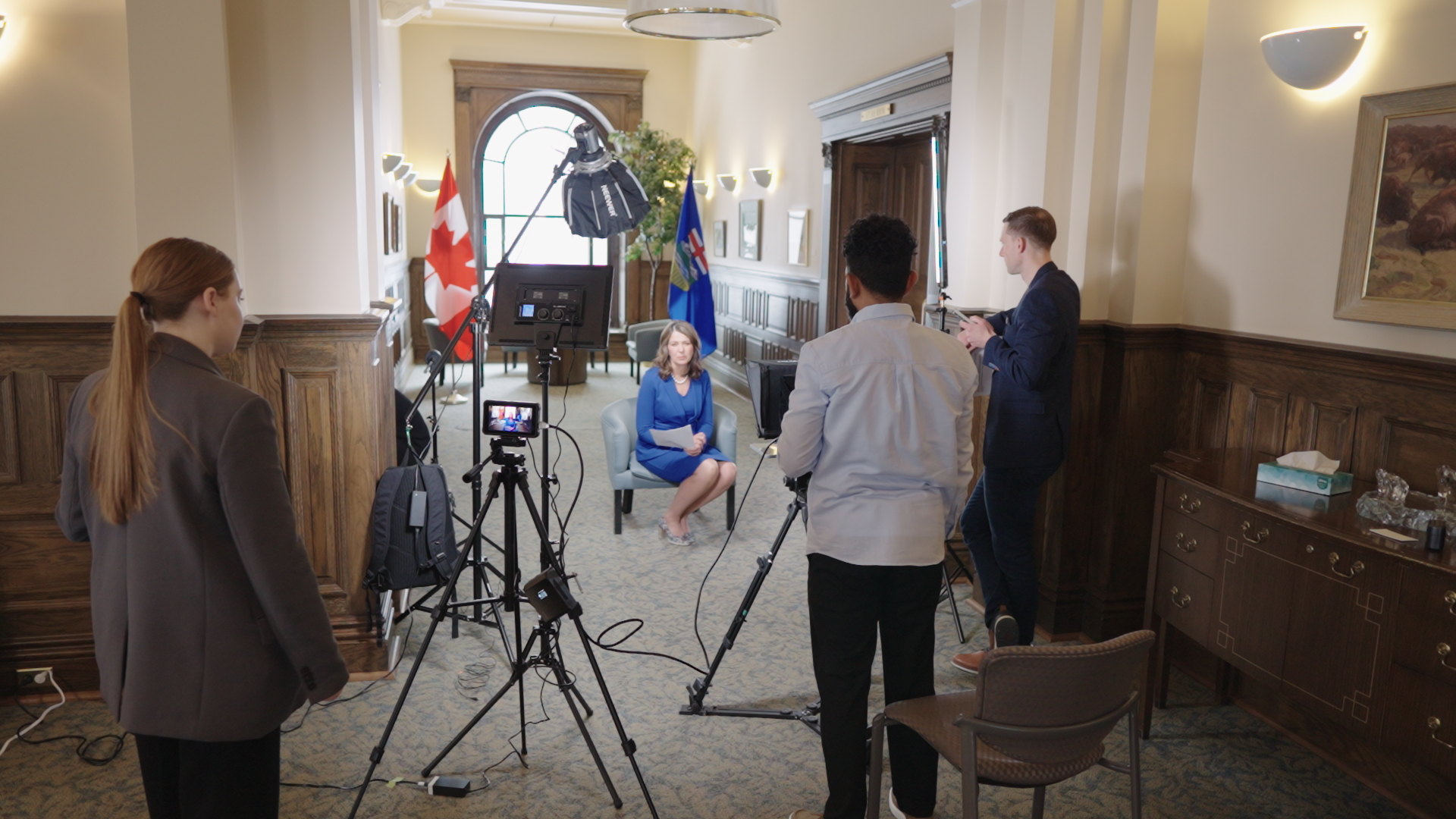 A woman in a blue dress being interviewed by a group of journalists in a room with wooden paneling and two flags behind her. There is professional filming equipment and lights set up.