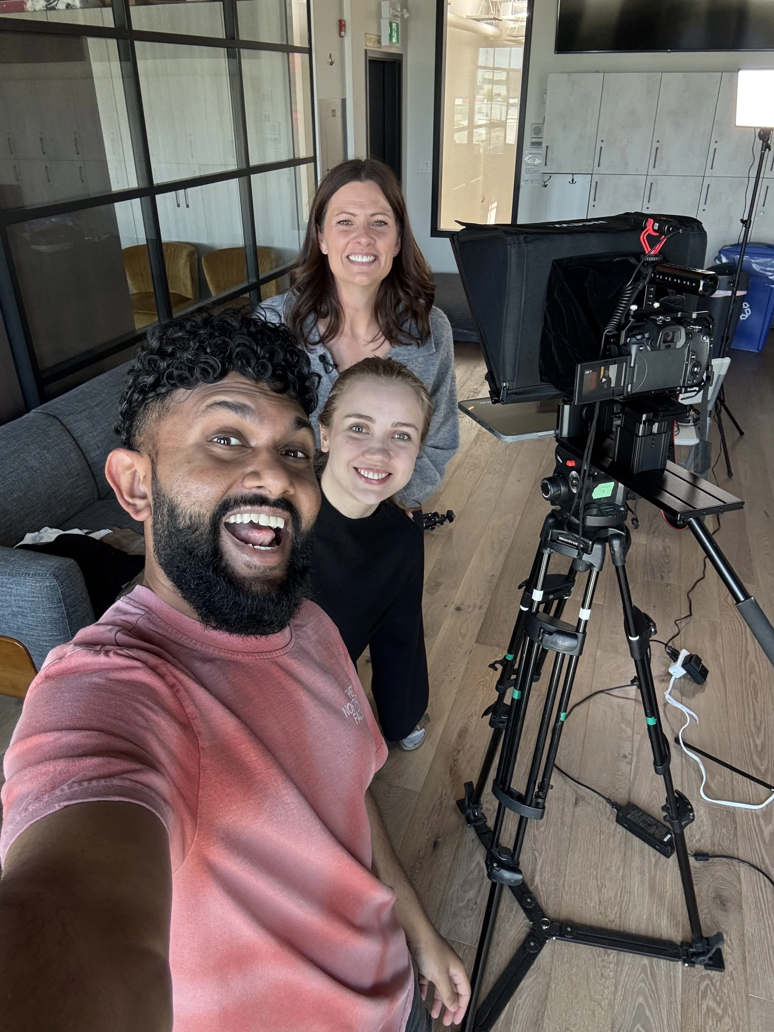Four smiling people taking a selfie in front of a professional video camera in a bright media room with wooden flooring and lockers.