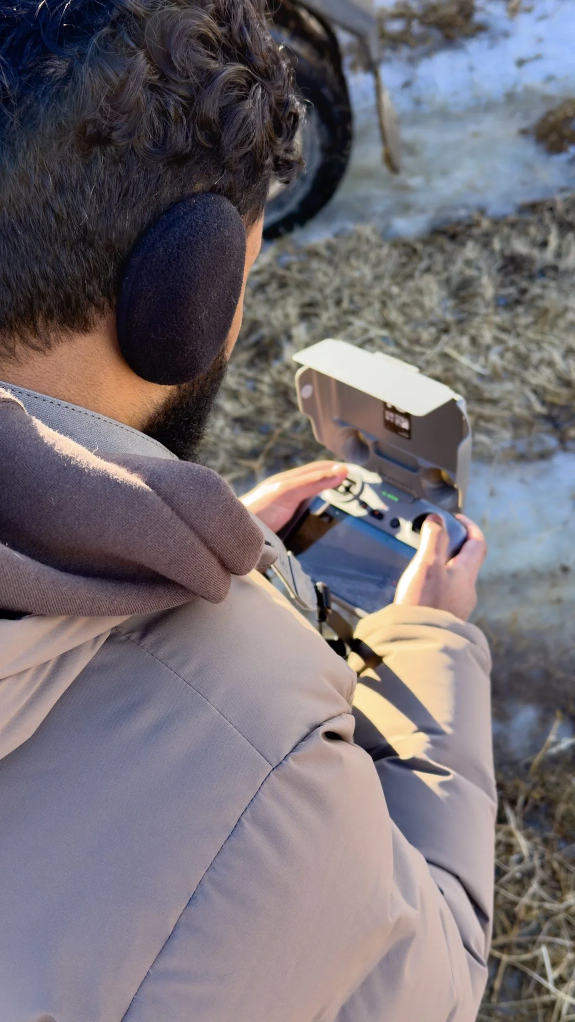 Person wearing beige jacket, with curly hair and black ear protection, operating a remote control drone outdoors near dried grass and water.