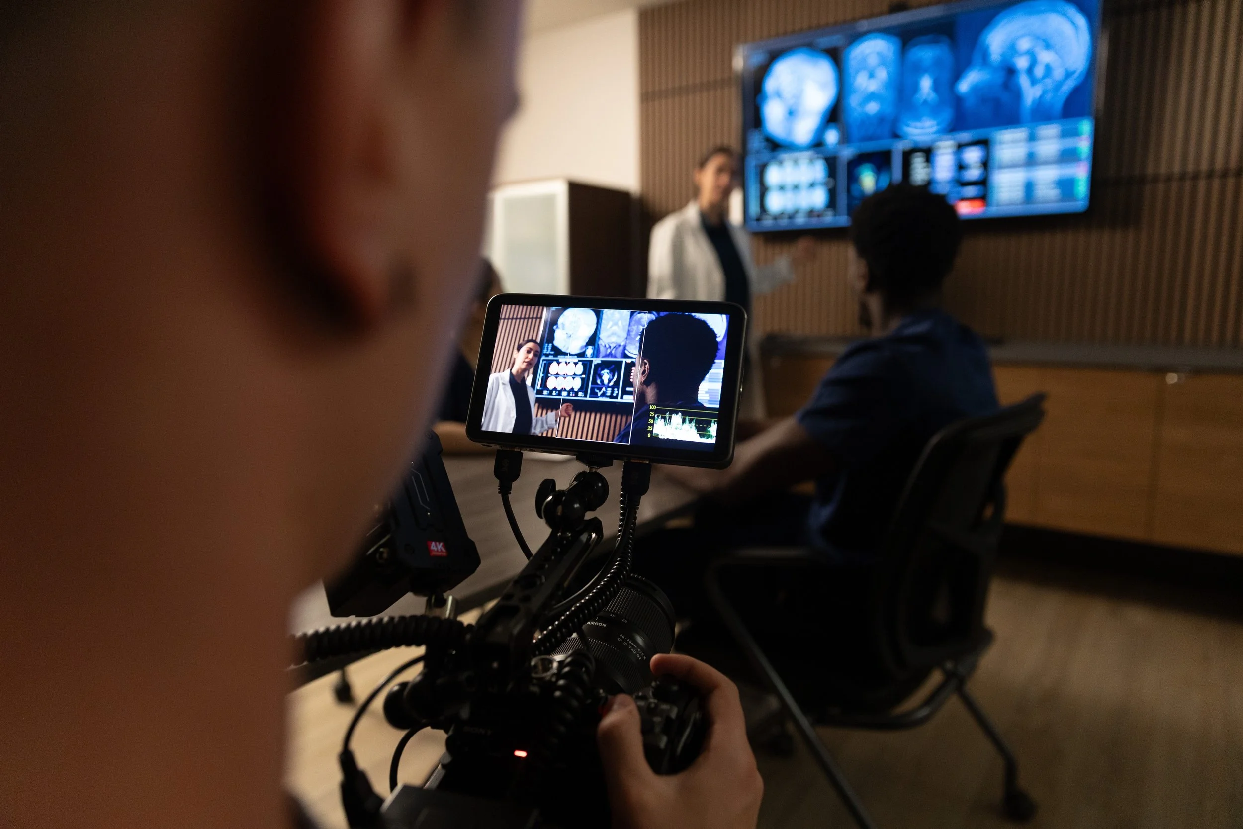 A person operating a video camera captures a scene in which a group of people, including a woman in a white coat, are seated and watching a presentation about medical scans displayed on a screen. The scene is set in a room with wood-paneled walls and