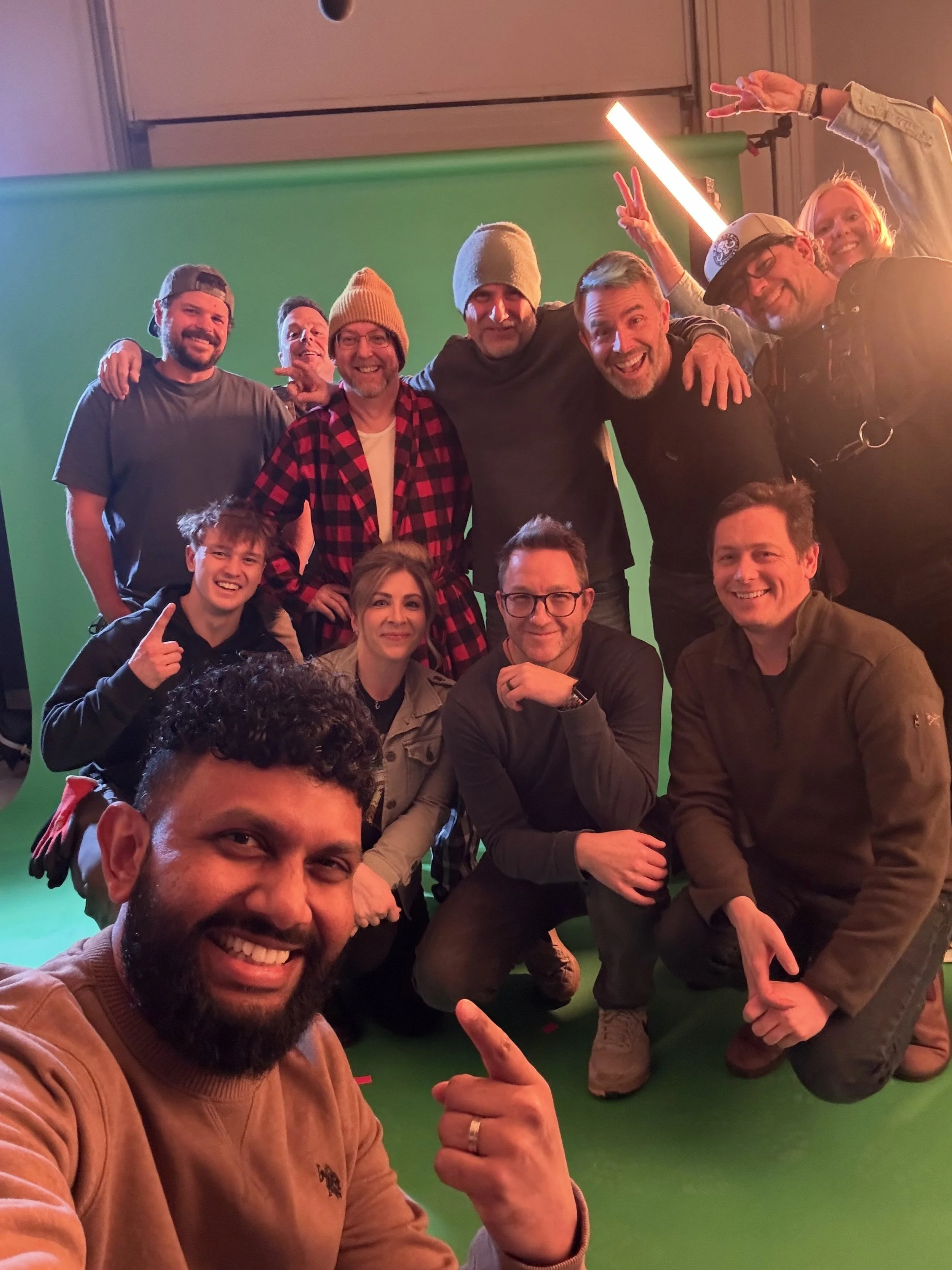 Group of people taking a selfie in front of a green screen, smiling and making peace signs.
