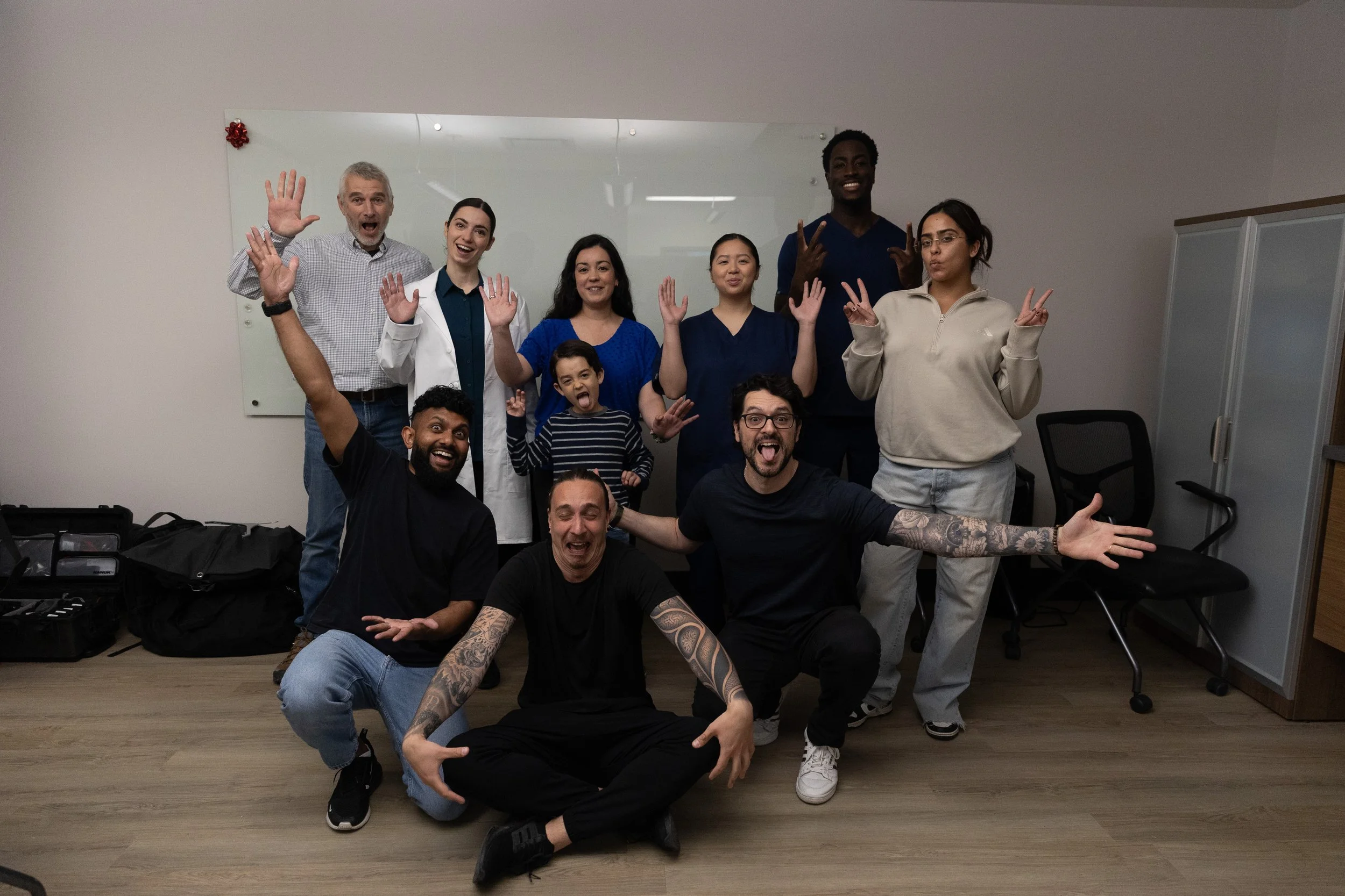 Group of nine diverse people posing playfully in an office room, some making peace signs and silly faces, with a whiteboard and lockers in the background.