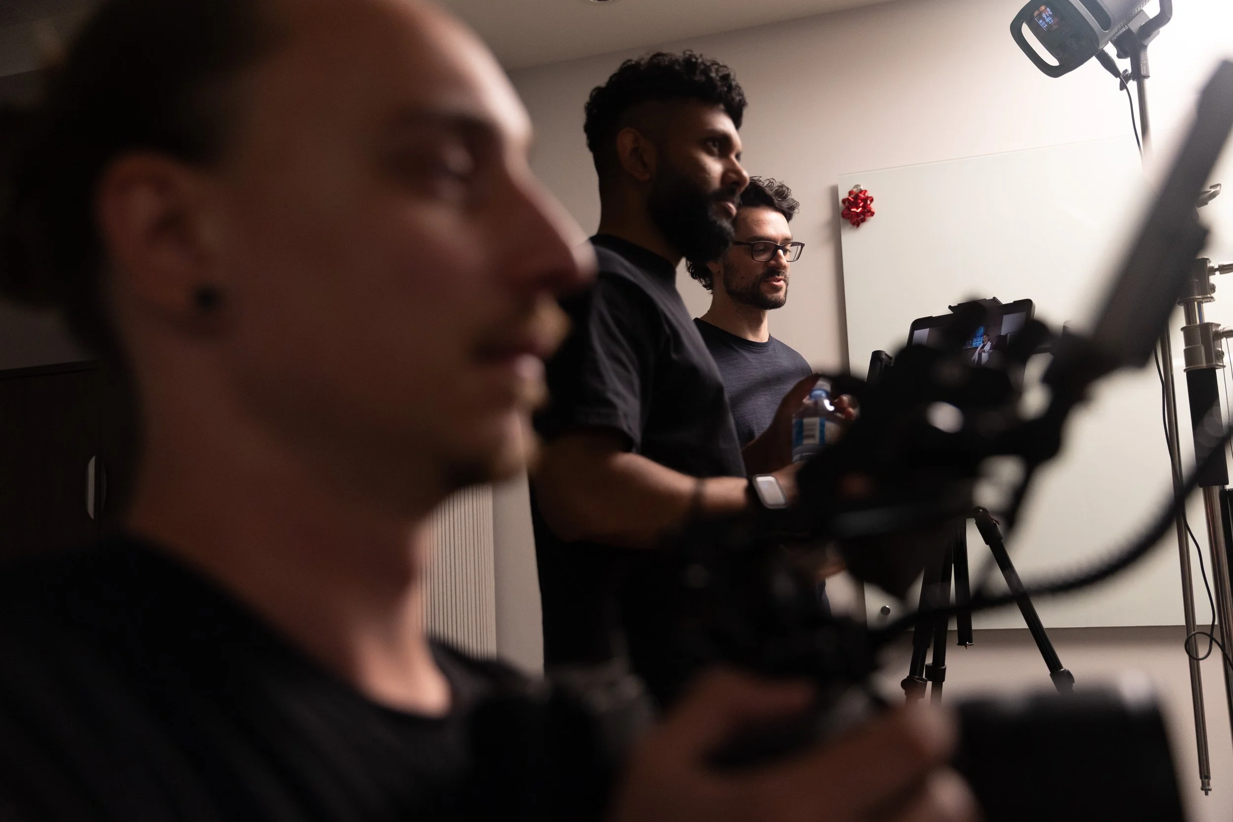Three men in black shirts watching a screen or camera in a room with minimal decor, including a white wall with a small red decorative item.