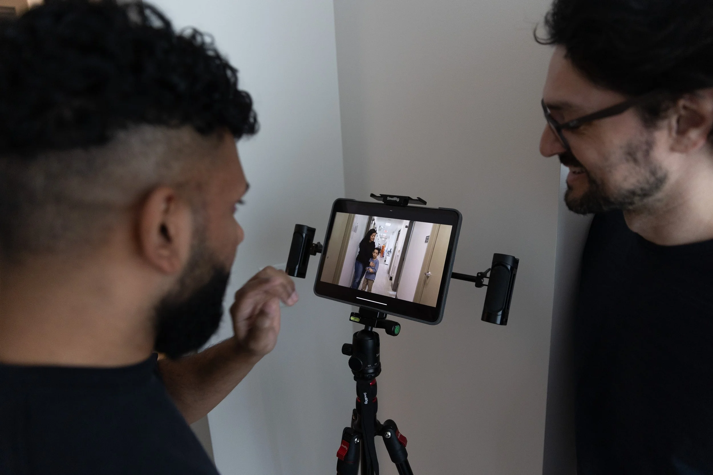 Two men are looking at a tablet on a tripod, capturing a photo of a woman and a child standing in a hallway. One man has glasses and a beard, the other has curly hair. The woman and child are visible on the tablet screen.
