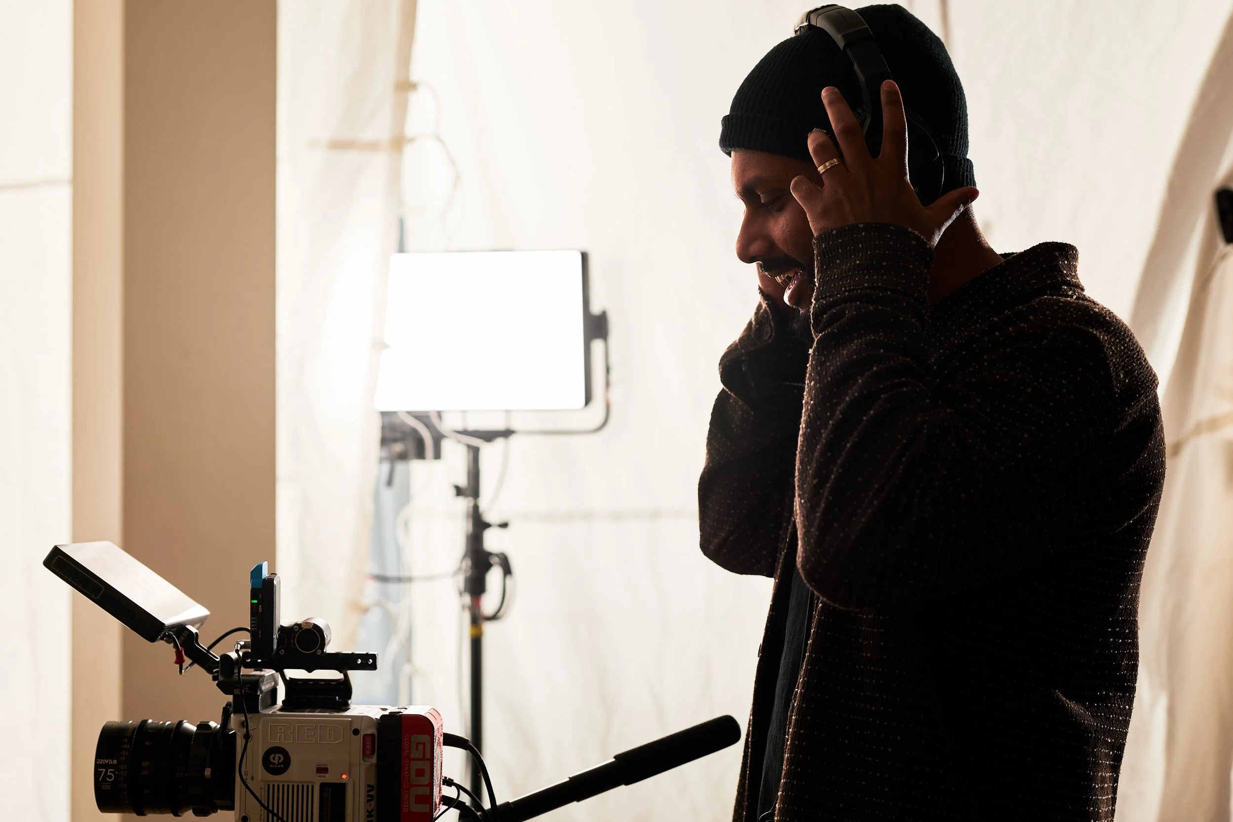 A person wearing a black beanie and dark sweater, smiling and holding headphones to their ear while working with camera equipment in a well-lit studio.