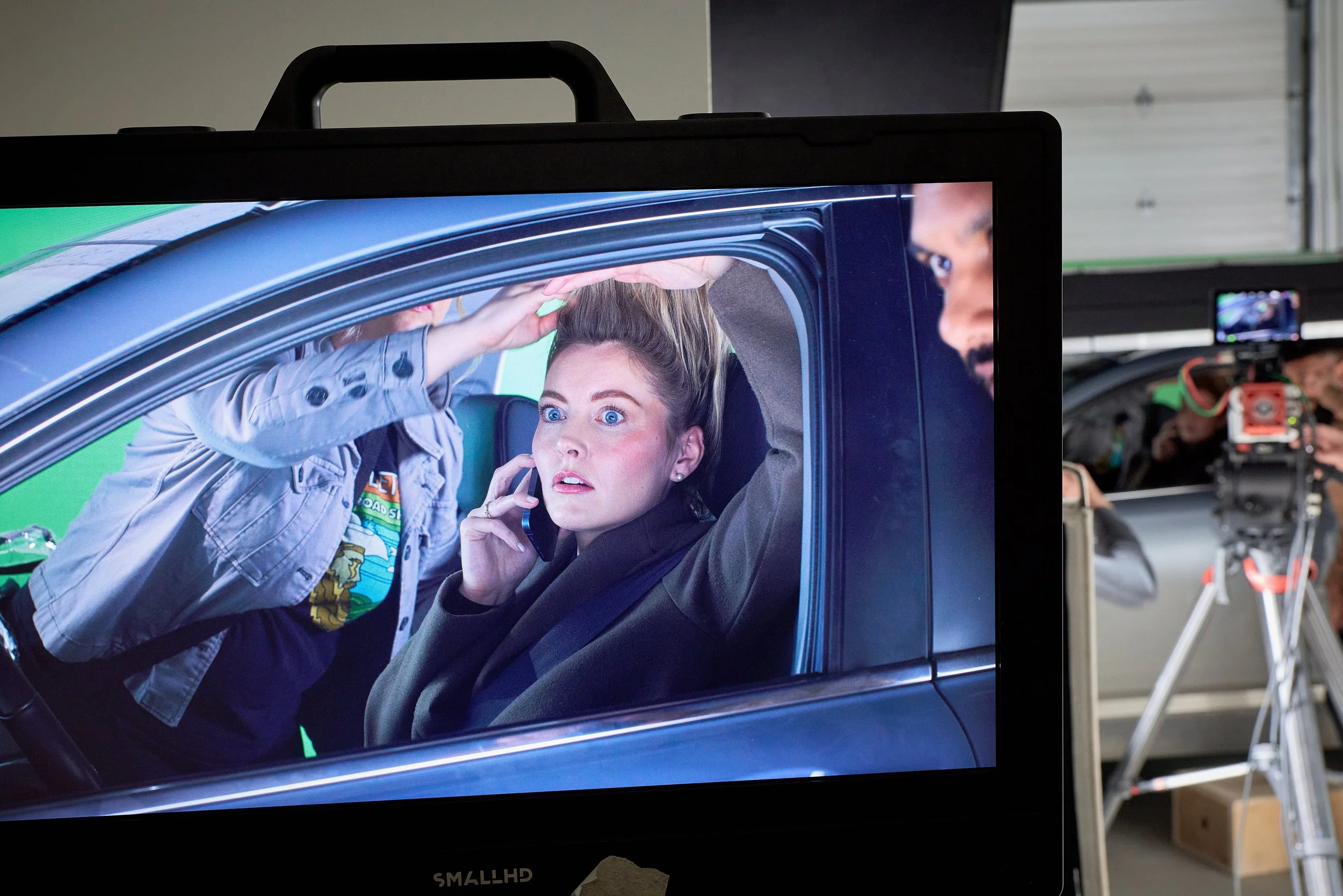 A woman with blue eyes and dark hair sitting inside a car, talking on a cell phone, and looking up with a surprised or curious expression. A person wearing a gray jacket with patches leans into the car through the window, touching the woman's head, w