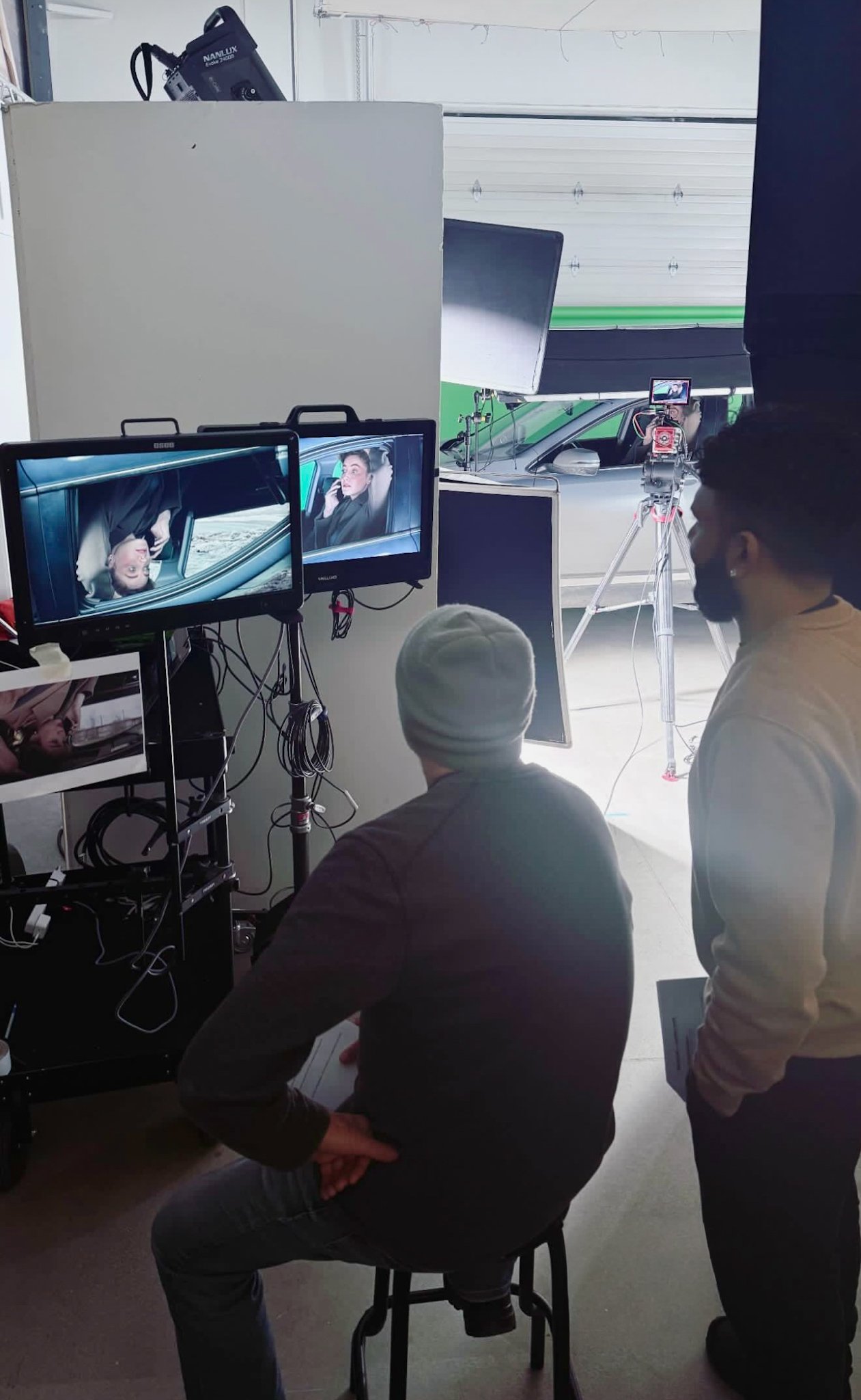 Two men are observing a video shoot setup with monitors showing a woman in a car speaking on the phone. One man is seated, and the other is standing, both looking at the monitors. The setup includes camera equipment, monitors, and lighting inside a g