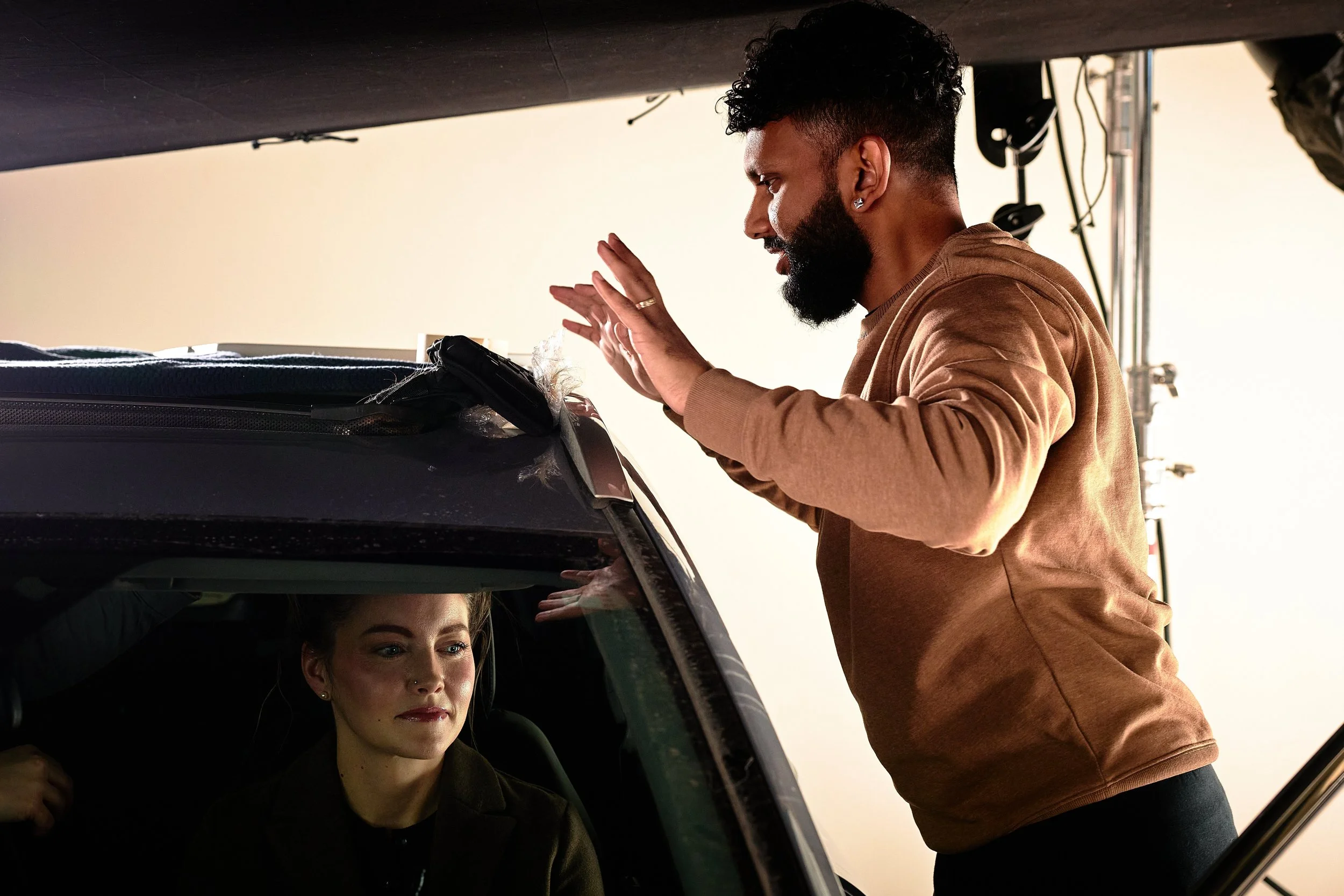 A man with curly hair and a beard leaning over the open roof of a car, talking to a woman sitting inside the car, with her face partially visible, in what appears to be a photo shoot setting.