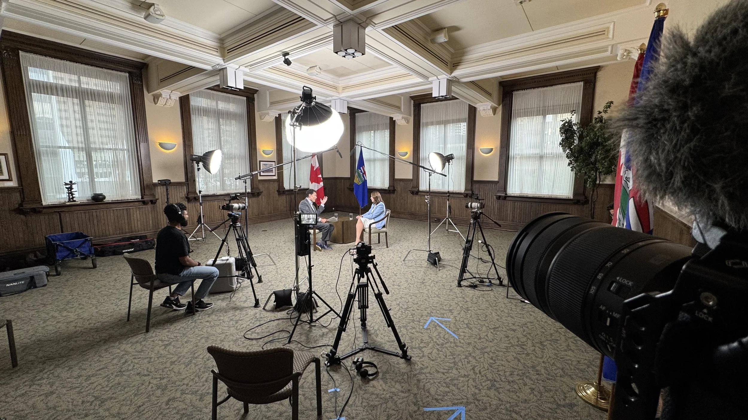 An indoor interview setting with two people seated and talking, Canadian and provincial flags in the background, surrounded by professional filming equipment including cameras, lights, and a backdrop.