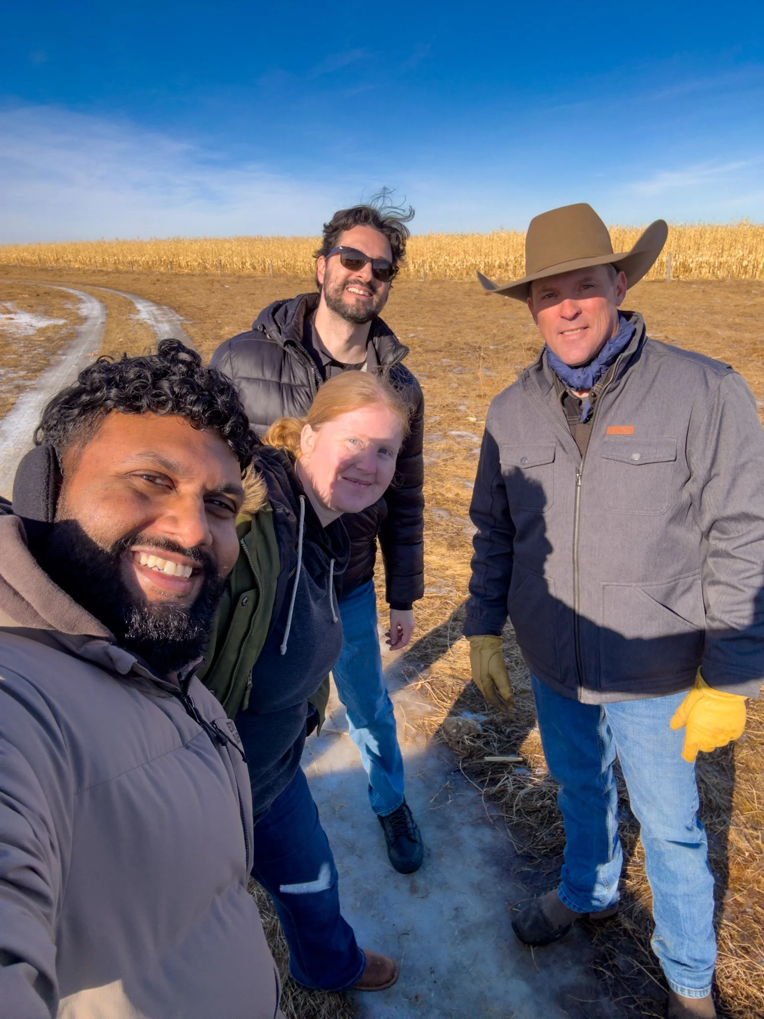 A group of five people taking a selfie outdoors on a sunny day with a field of corn and a clear blue sky in the background.