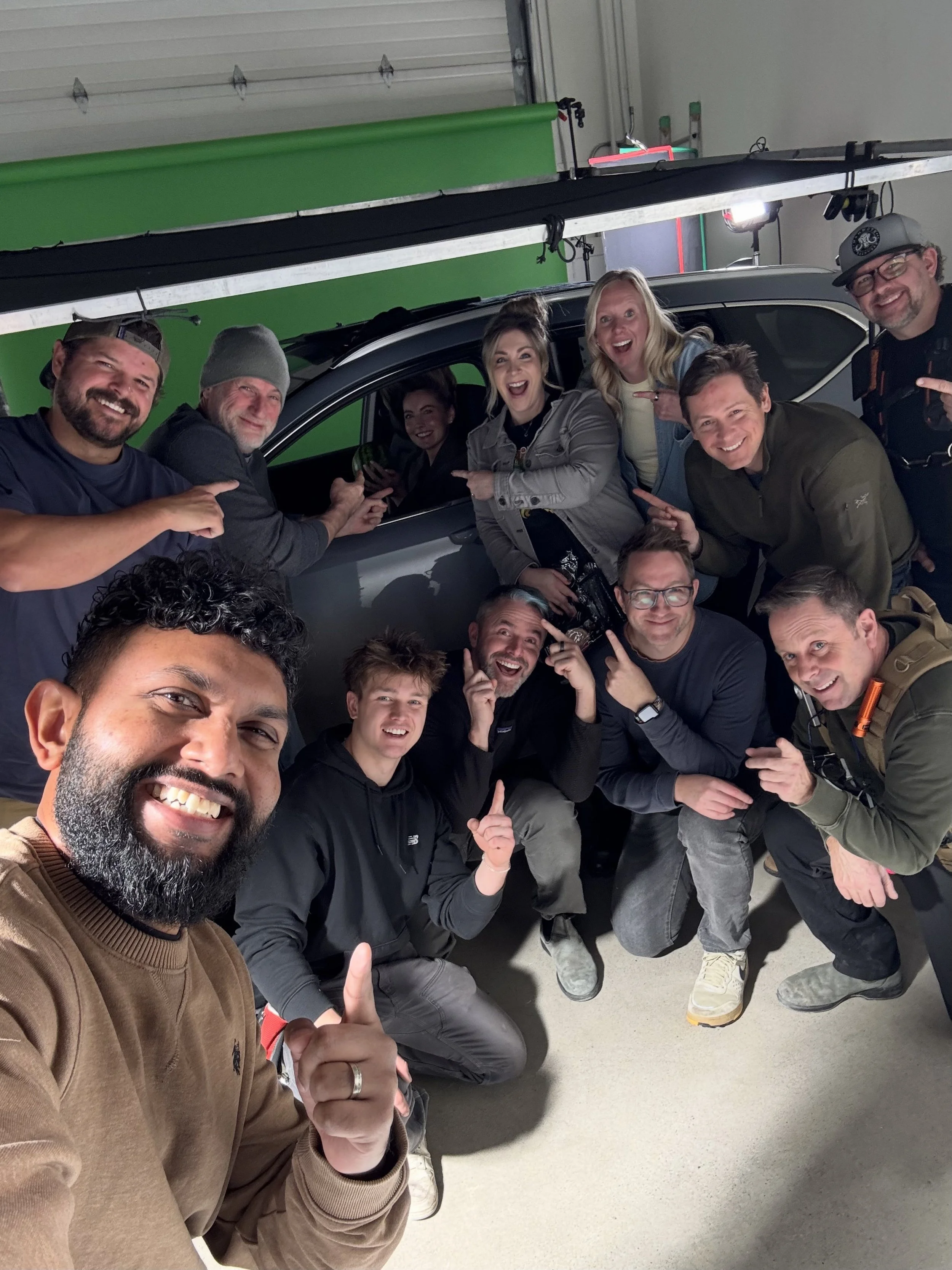 A group of people smiling and pointing at the camera inside a garage with a car and a green screen backdrop.