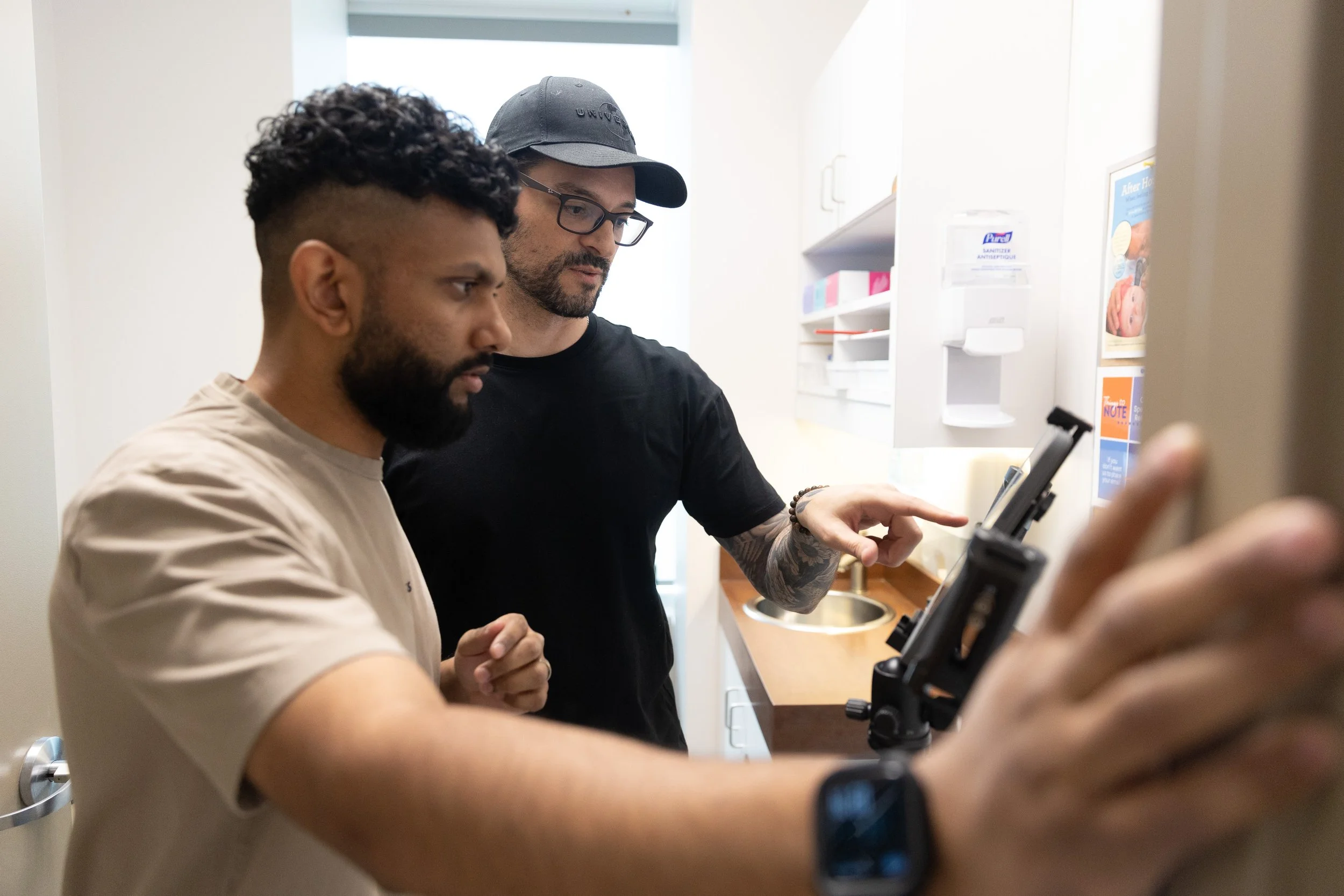 Two men looking at a tablet in a kitchen, one with a beard and the other with glasses and a baseball cap.