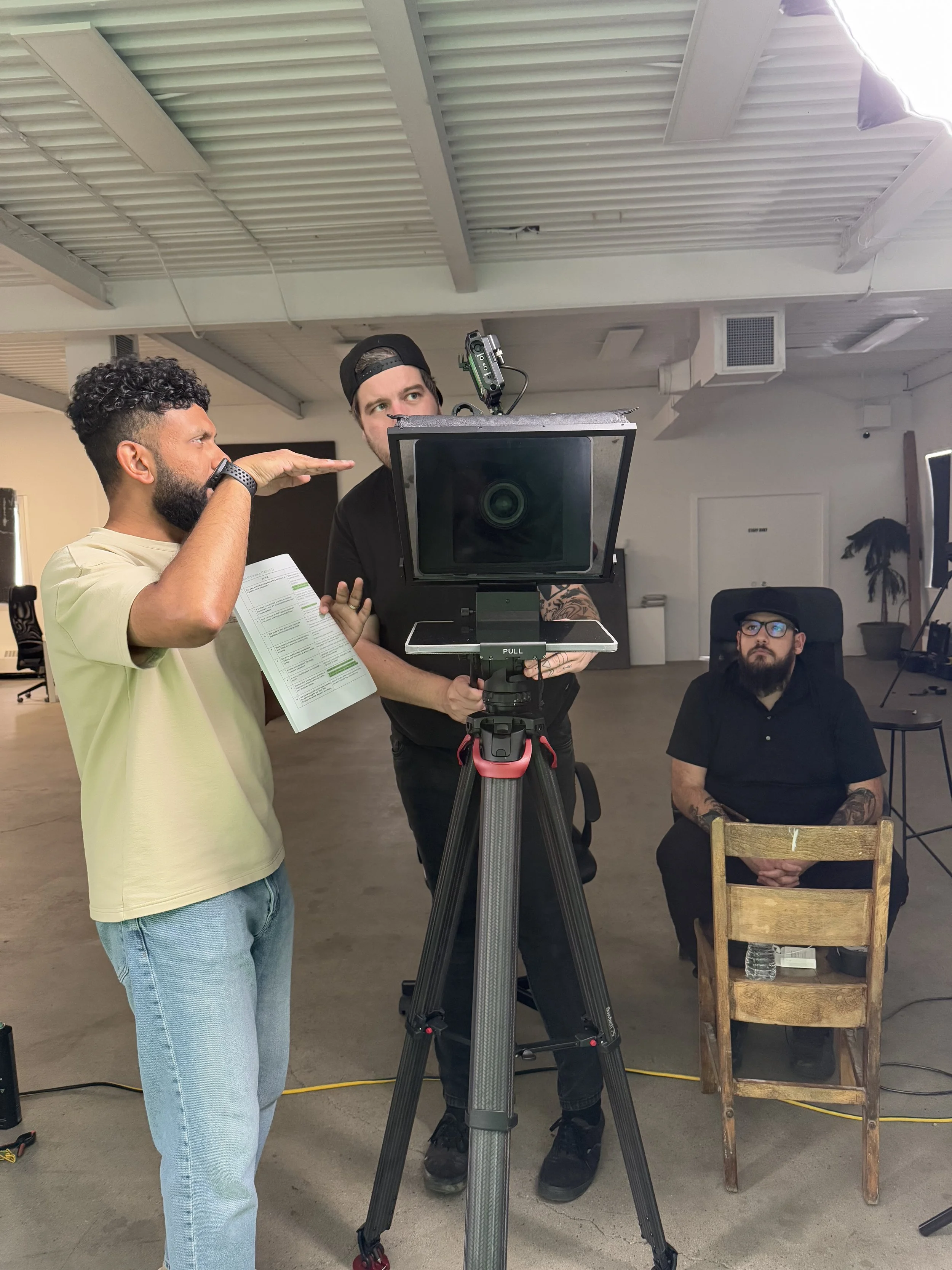 Three men in an industrial space working with filming equipment. One man is standing and gesturing towards a screen, another is operating a camera on a tripod, and the third is sitting on a wooden chair with tattoos on his arms, wearing glasses.