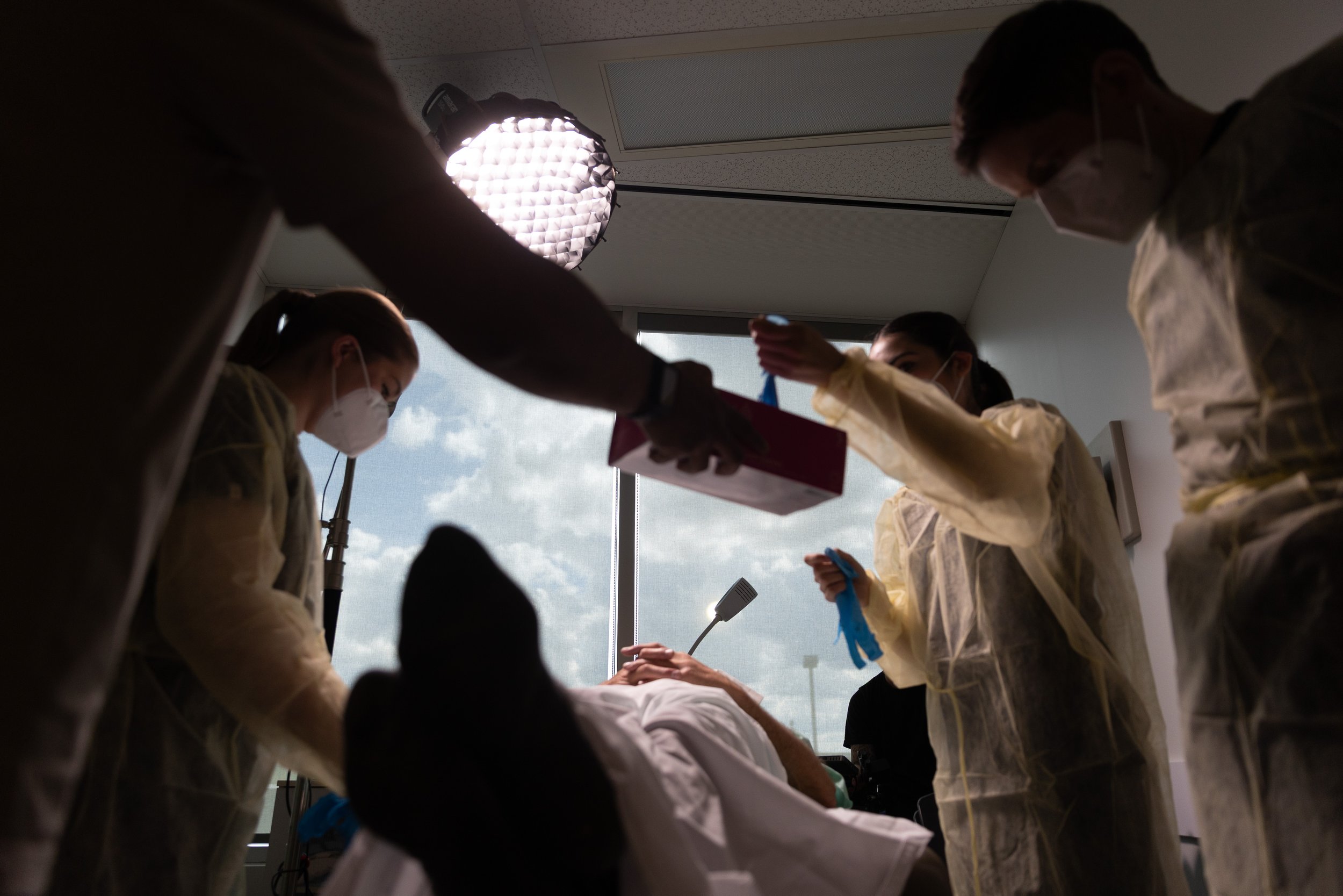 Medical professionals in protective gowns, gloves, and masks working together in a hospital room, with natural light coming through a window.