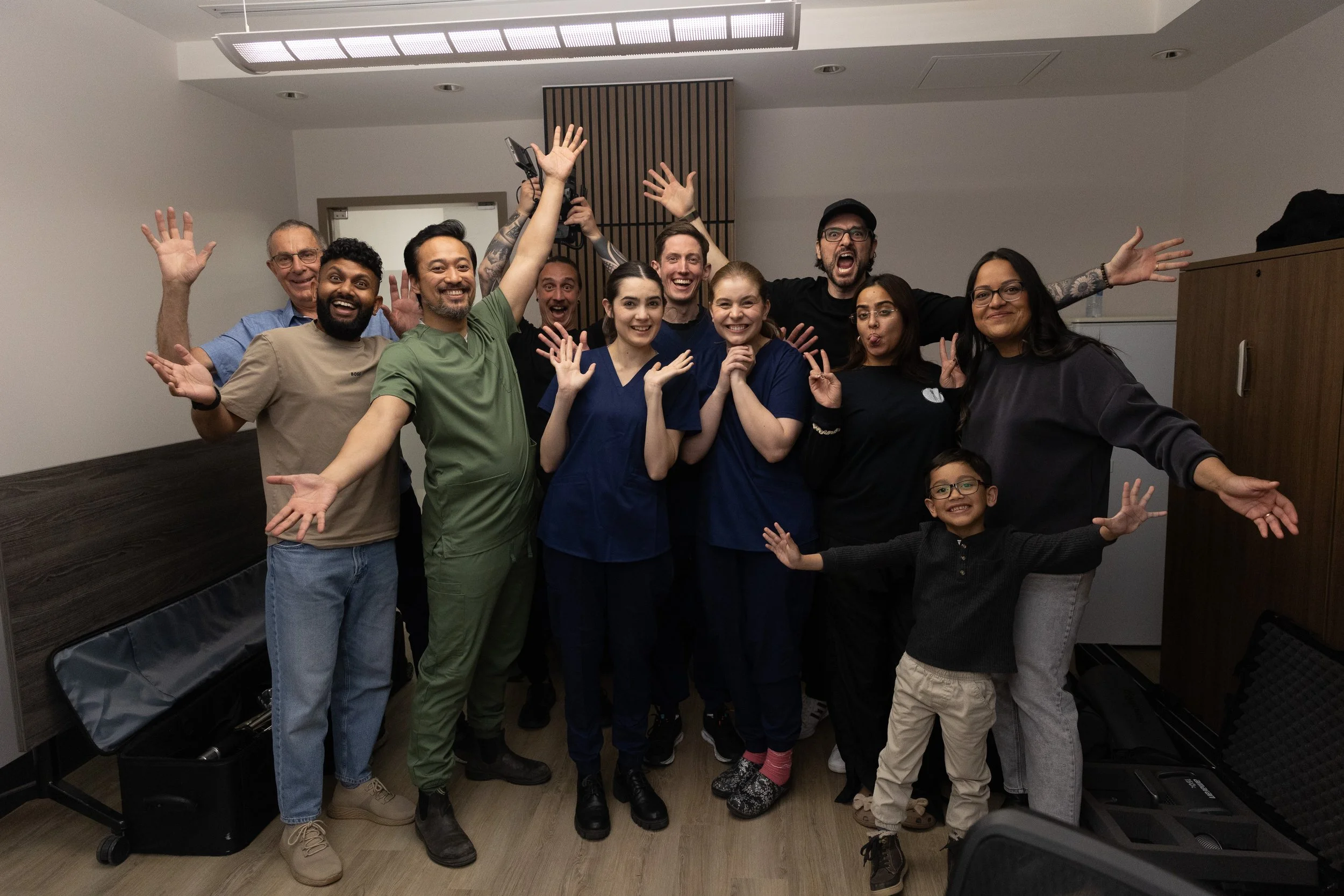 Group of eleven diverse people smiling, waving, and posing for a photo in an indoor room.