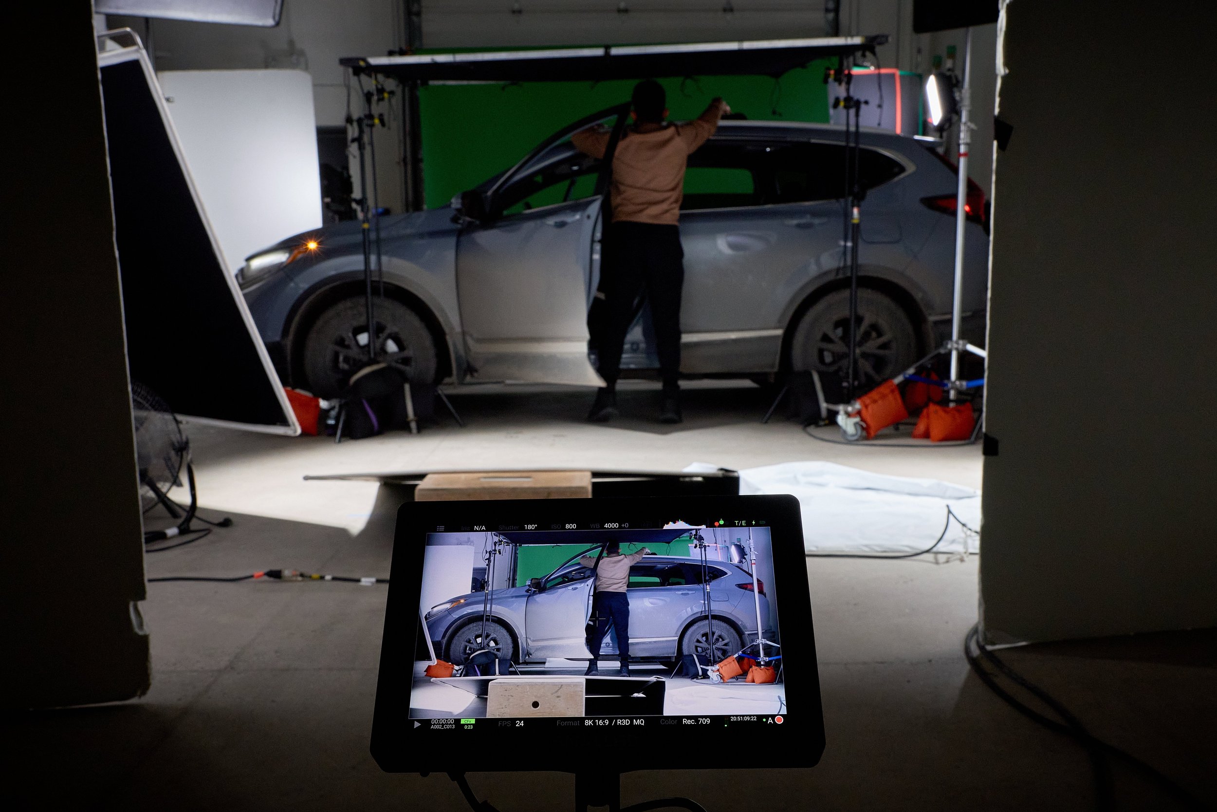 A behind-the-scenes shot of a video shoot featuring a gray car in front of a green screen, with a man adjusting equipment and a monitor displaying the scene being filmed.