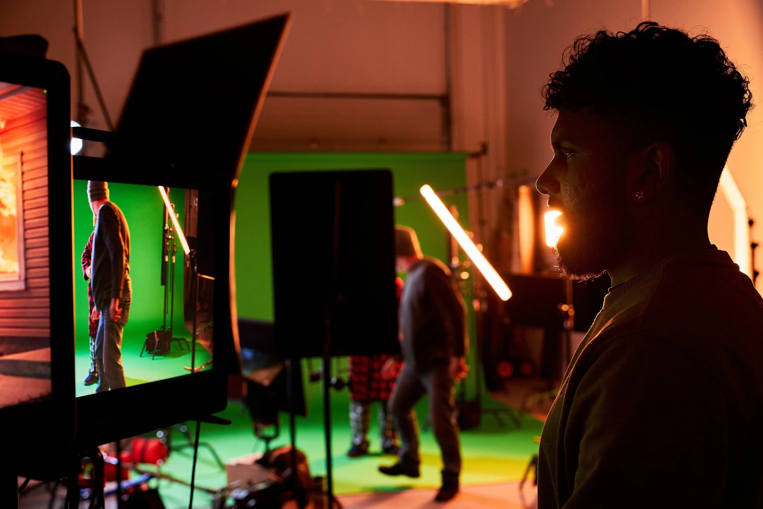 A person with curly hair is observing a scene with a green screen and filming equipment, including a monitor displaying the scene and crew members working, inside a dimly lit studio.