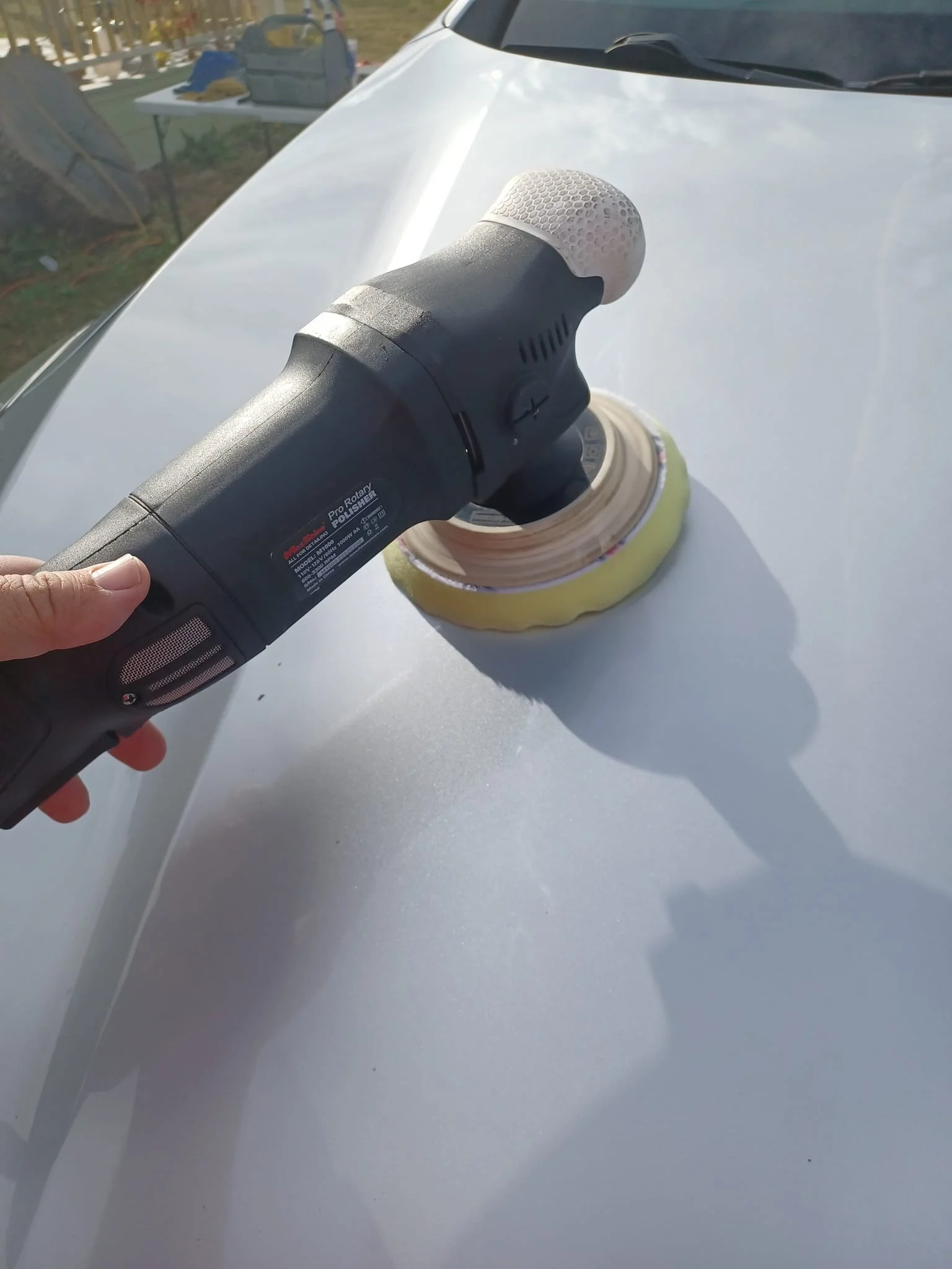 Person using a rotary buffer to polish a white car hood outdoors.