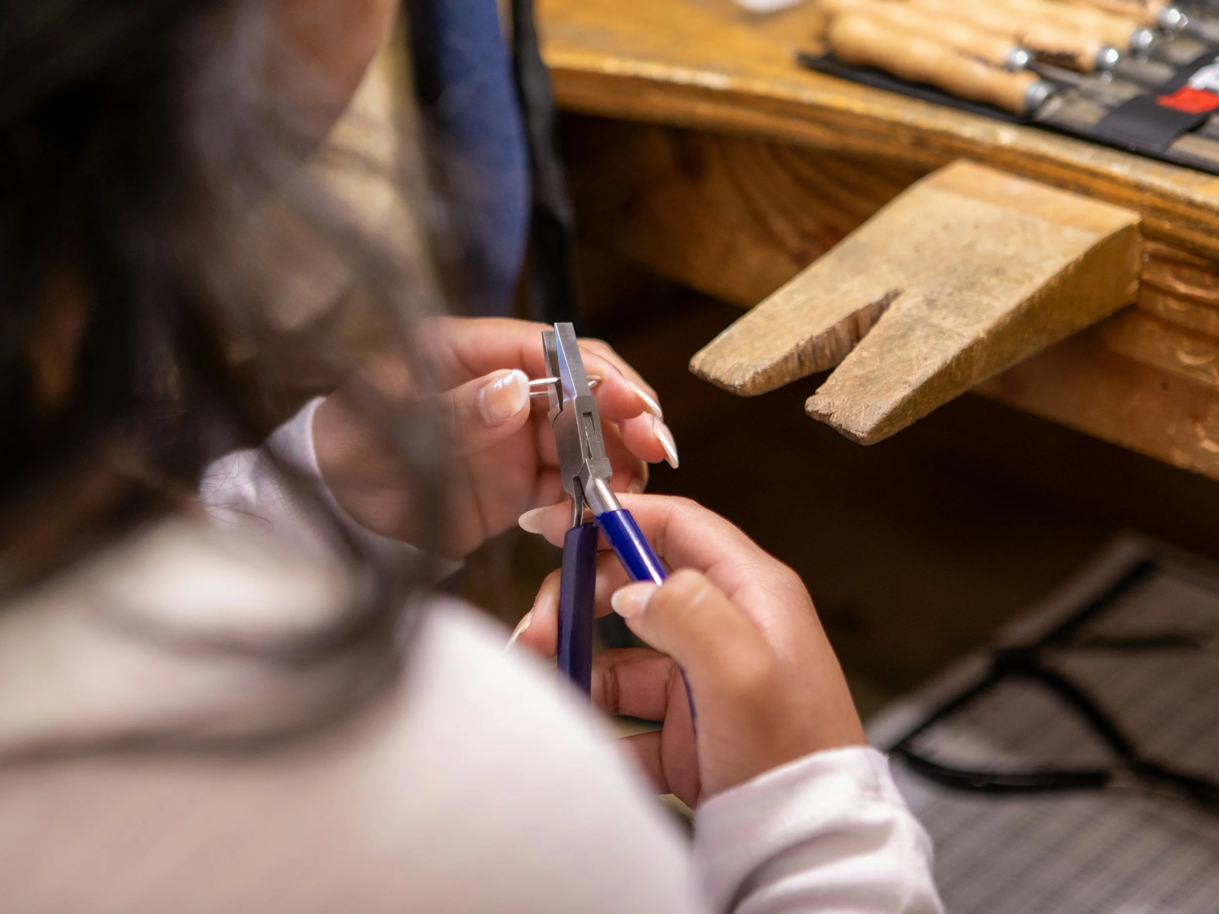 A person using jewelry pliers to work on a small piece of jewelry at a workbench with woodworking tools.