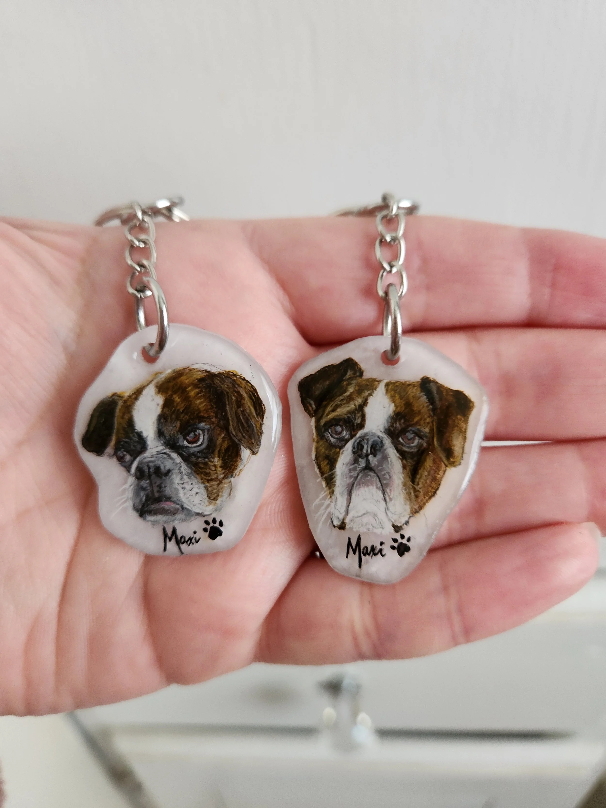 Hand holding a pair of dog-themed earrings with painted images of a dog’s face, the name 'Maxi' and a paw print below.